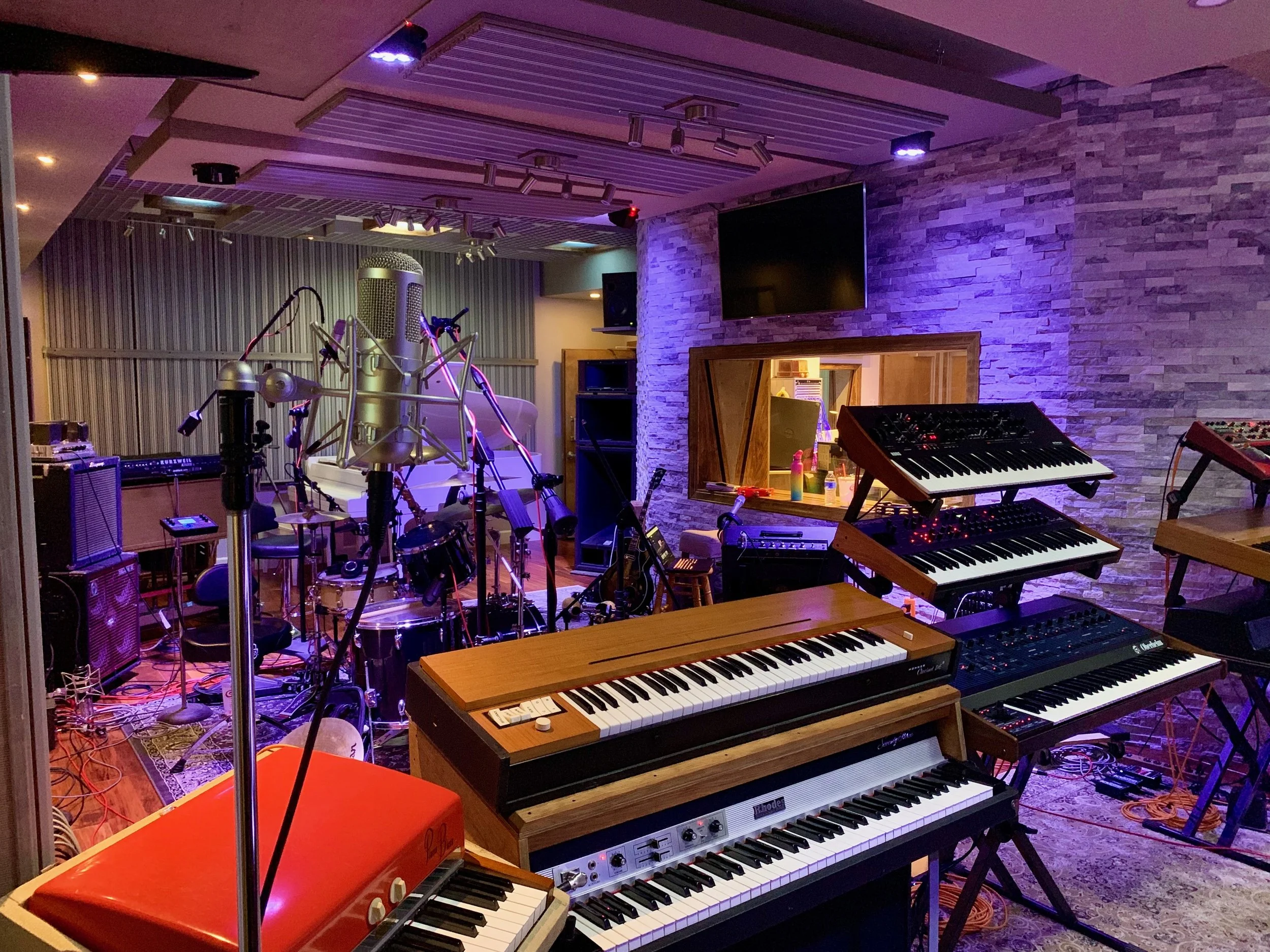 A recording studio with multiple musical instruments including Korg and Roland keyboards, a drum set, microphones, and audio equipment, with purple and warm lighting and a stone wall backdrop.