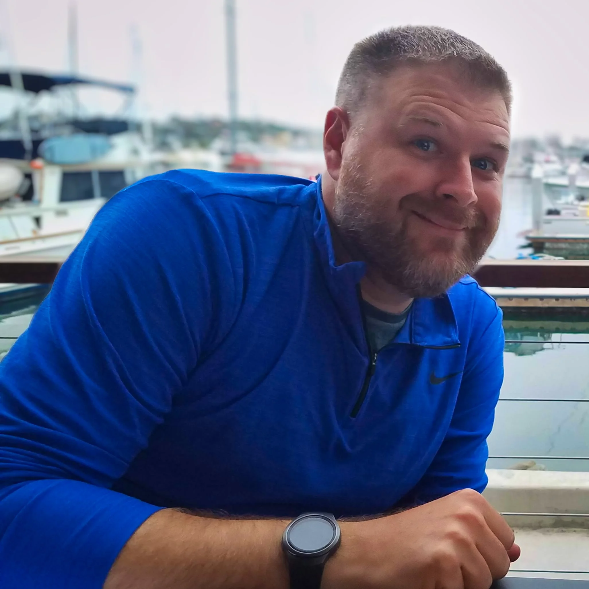 A person with short hair and a beard leaning on a table wearing a blue jacket and a watch, with boats docked in the background near a body of water.