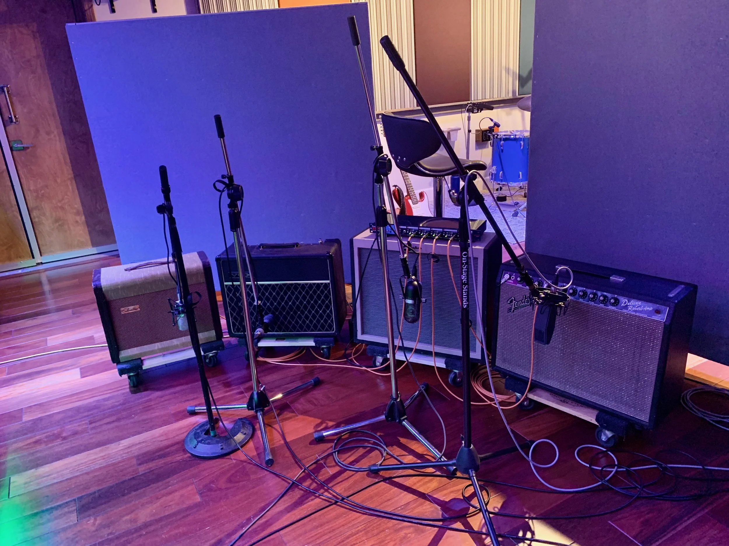 Music studio setup with guitar amplifiers, microphone stands, acoustic panels, and musical instruments including a guitar and a drum set in the background.