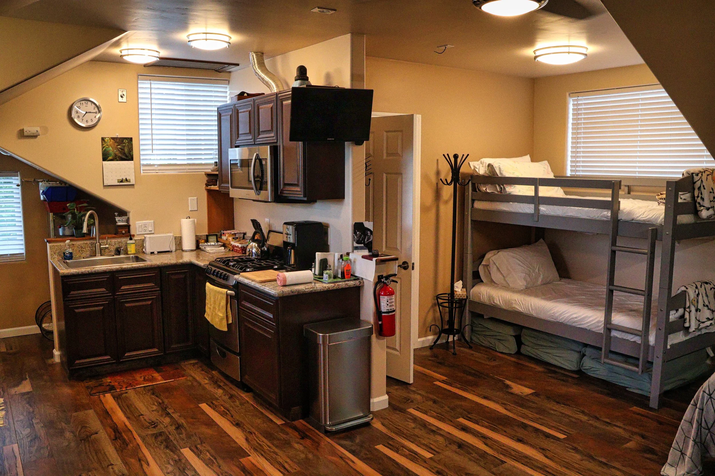 Interior view of a small living space with a kitchen and sleeping area. The kitchen has dark cabinetry, a granite countertop, a stainless steel sink, and appliances including a microwave and stove. A flat-screen TV is mounted on the wall above the st