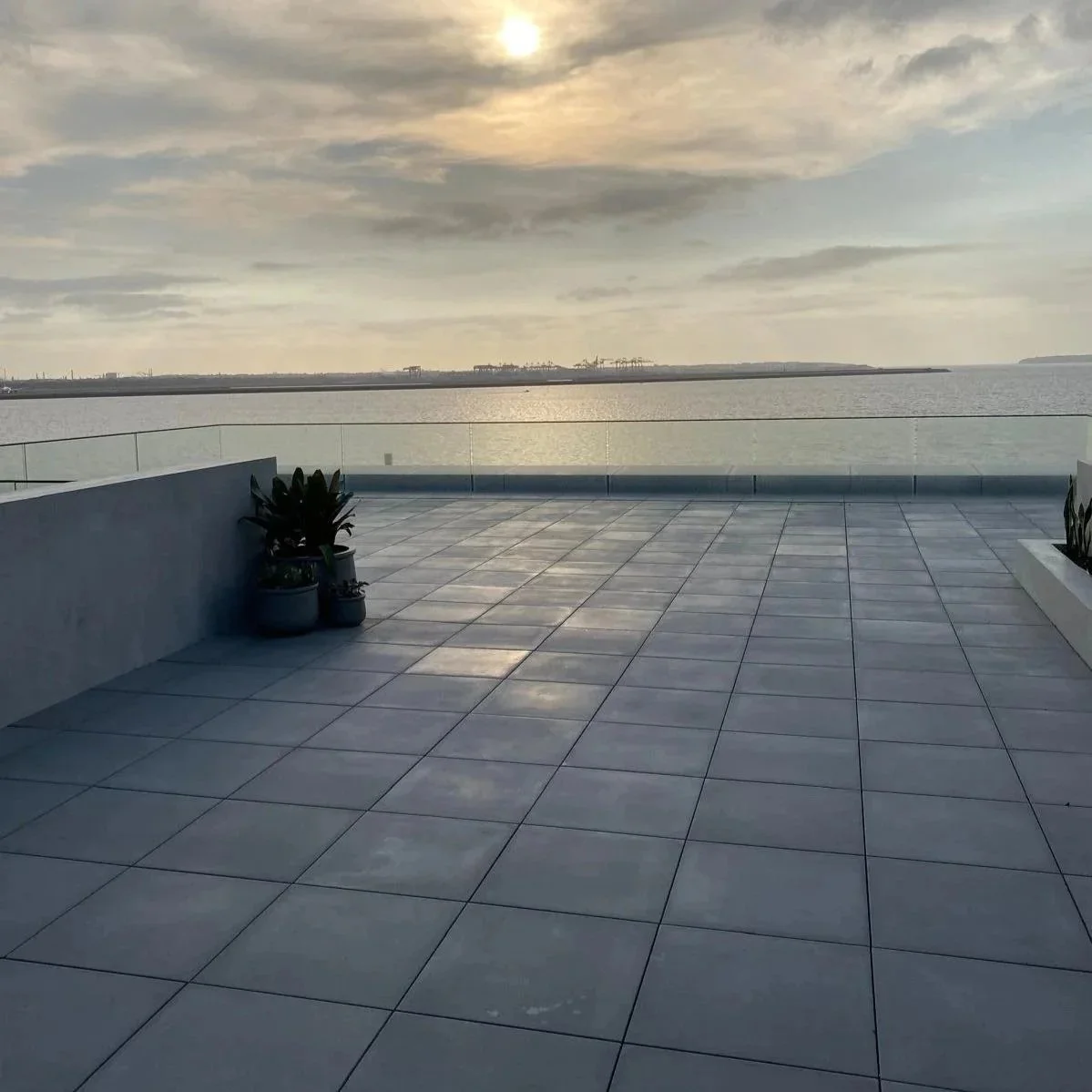 An outdoor terrace with large white tiles, potted plants in the corner, glass railing, overlooking a body of water with an industrial area in the distance under a cloudy sky with the sun partially visible.