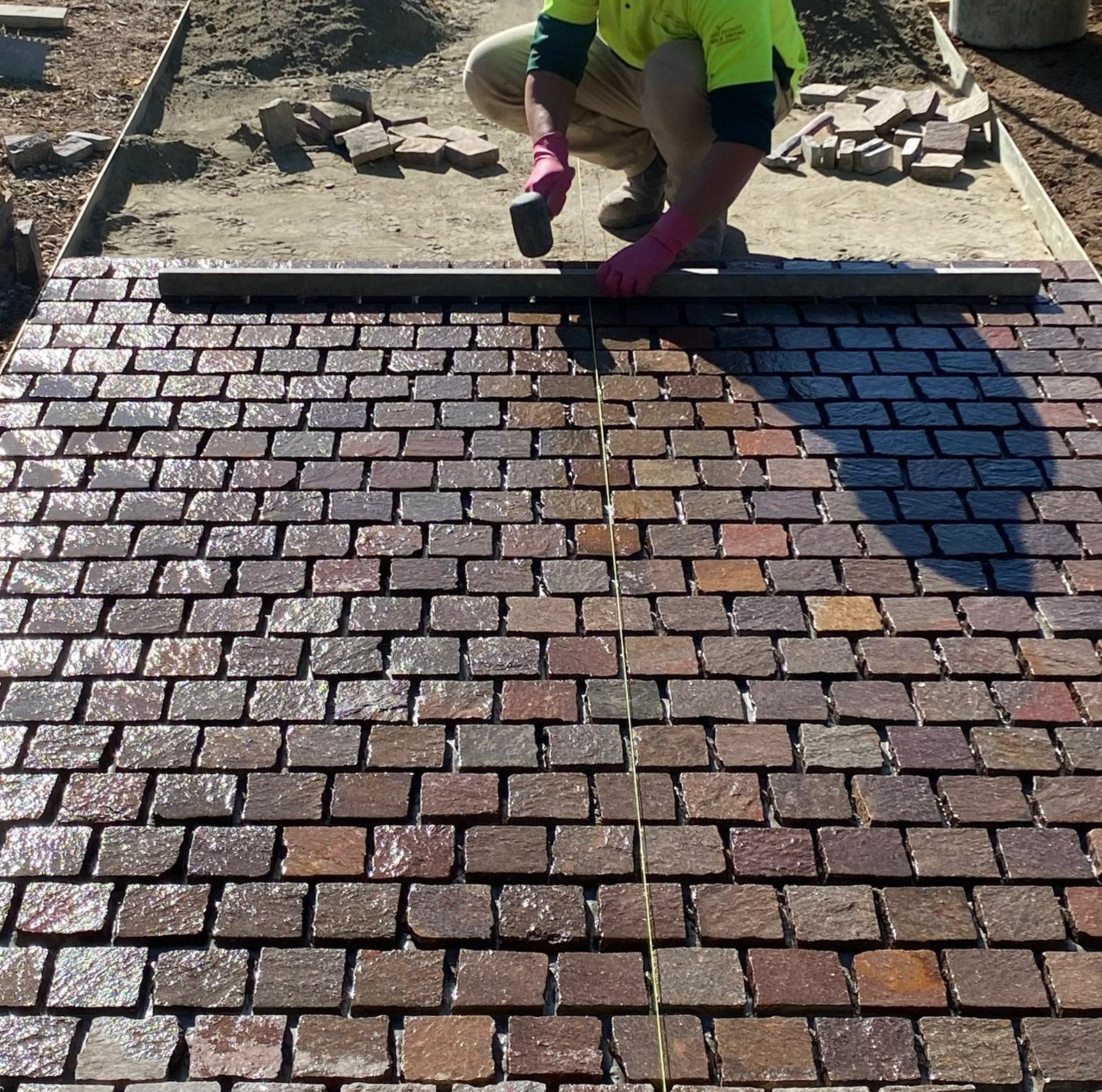 A person laying bricks on a newly paved walkway or driveway, wearing pink gloves and a bright yellow safety vest, with a measuring tape stretched out along the surface.