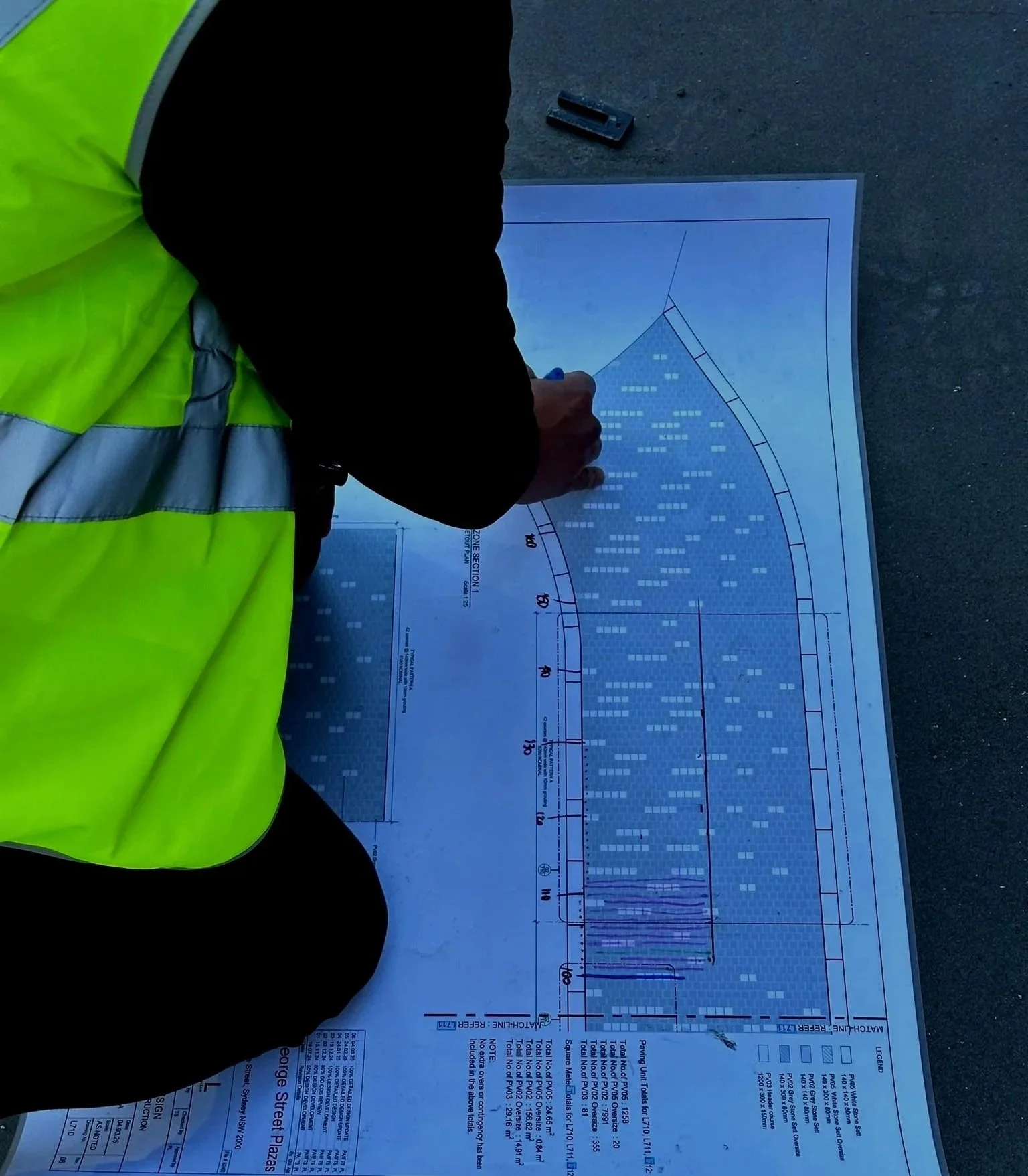 Person in a yellow safety vest marking a detailed construction or engineering plan on the ground.