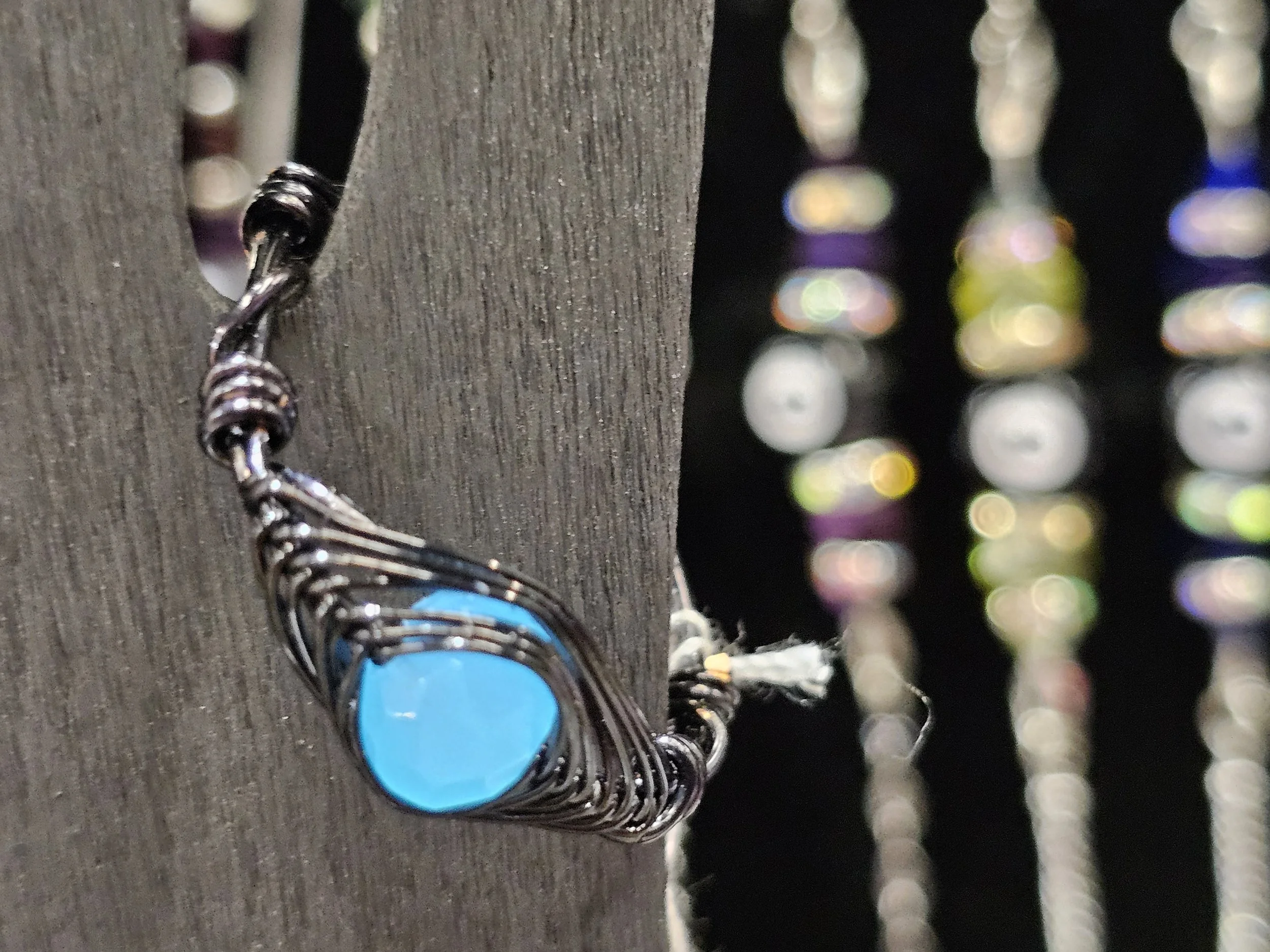 Close-up of a blue pendant necklace hanging on a gray string with a blurred background of hanging beads or chains.