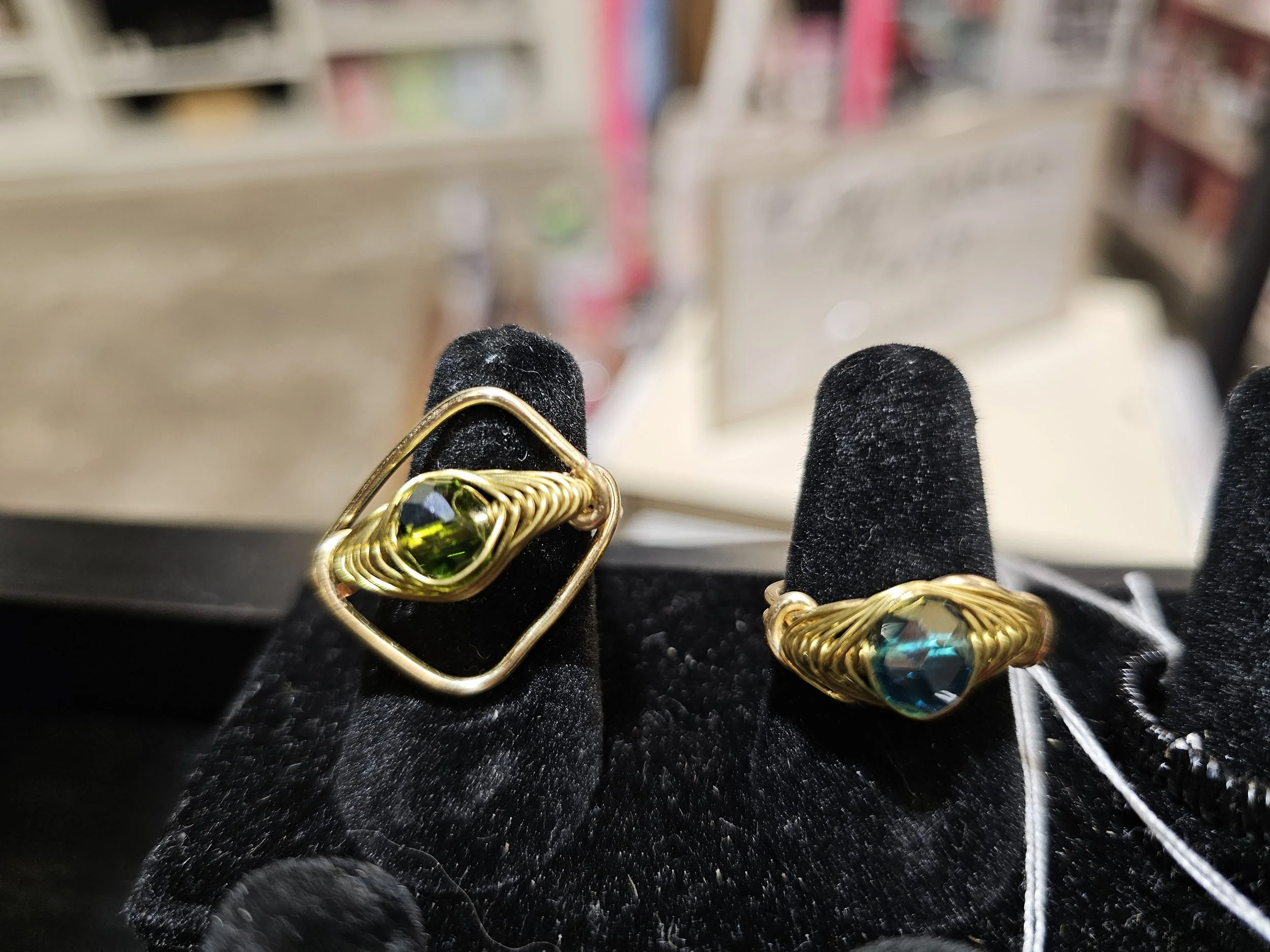 Two gold rings with large, colorful gemstones displayed on black velvet surface. One ring has a greenish gemstone, and the other has a bluish gemstone, both set inside intricate wire wrapping.