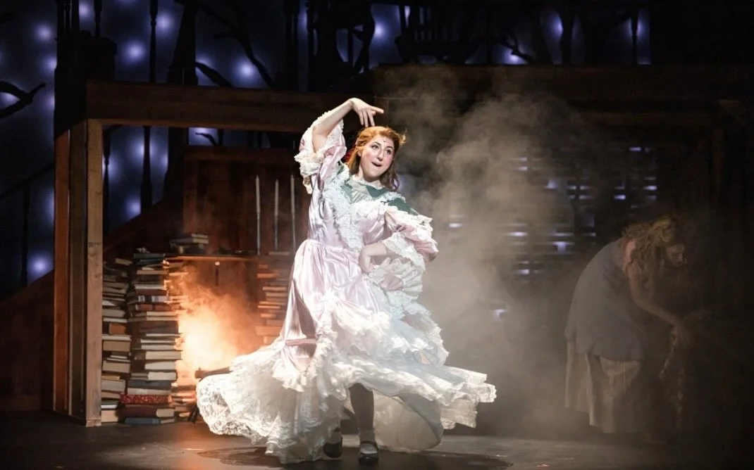 A woman in a flowing, pale pink dress performing on stage with books and smoke behind her, and a person in the background at a piano.