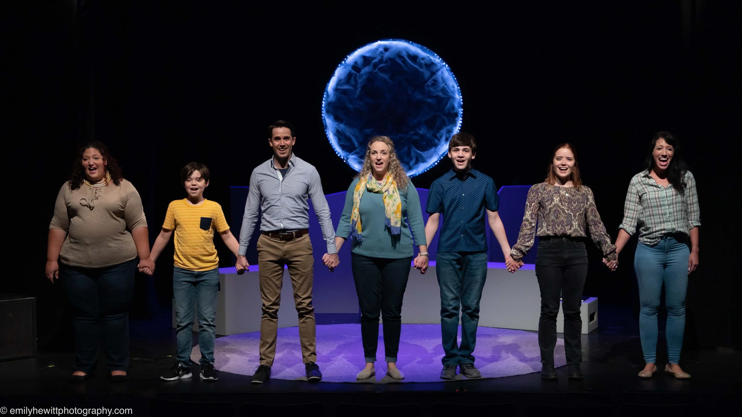 People holding hands on a stage in front of a dark backdrop with a lit circular prop behind them.