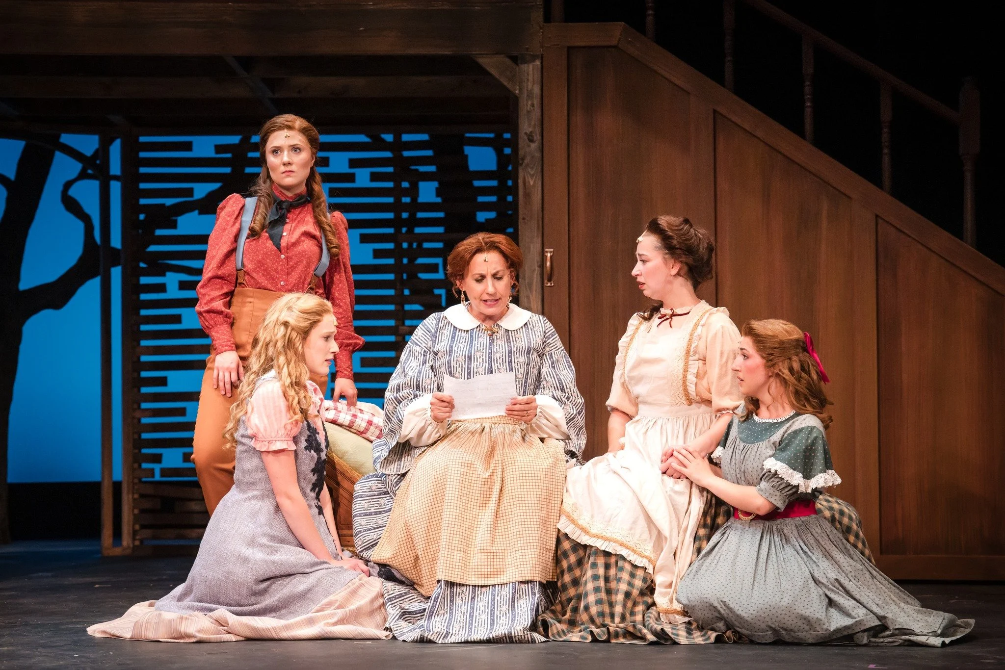 Five women dressed in period costumes on stage, one reading a letter while the others listen, set against a wooden interior and a cutout background of a tree.