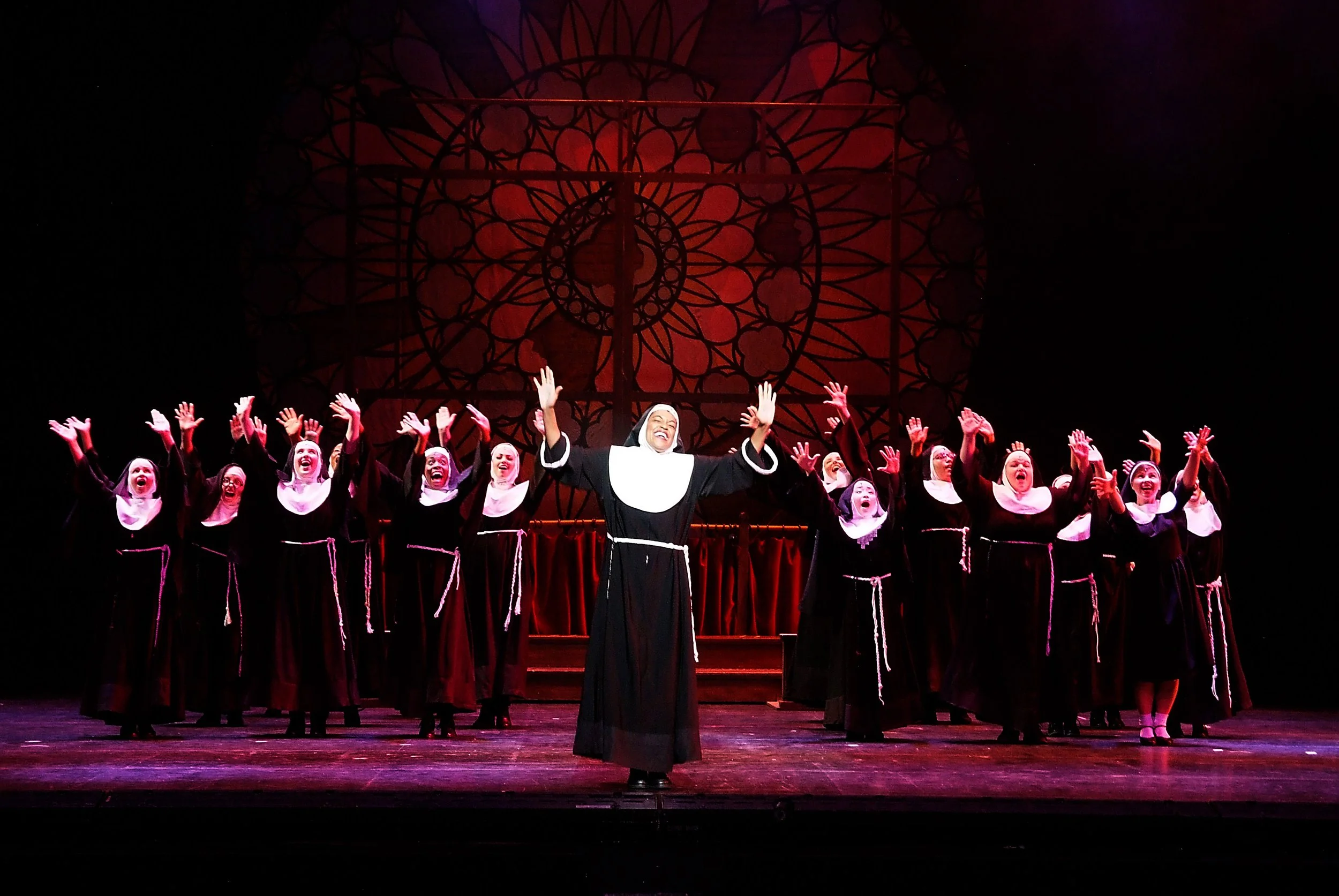 Women dressed as nuns performing on stage with arms raised and smiling, background features a large decorative stained glass window.