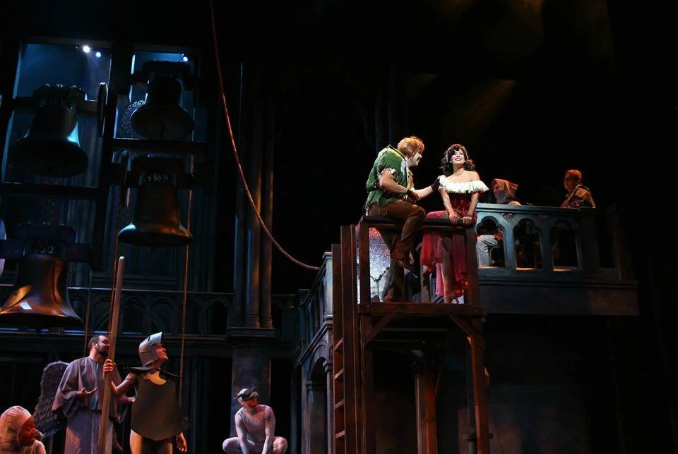 Actors on stage performing in a theater, with some on a balcony and others below, surrounded by large hanging bells.