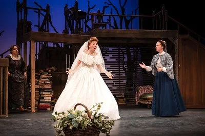 Three women performing on stage, one in a white wedding dress, likely acting in a play or musical, with a set resembling a rustic home background.