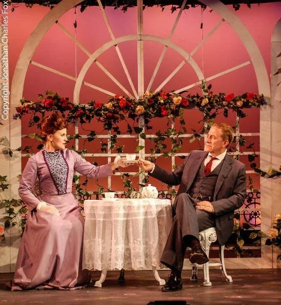 Two actors on stage, a woman and a man, sitting at a table with a floral backdrop, engaging in a theatrical scene.