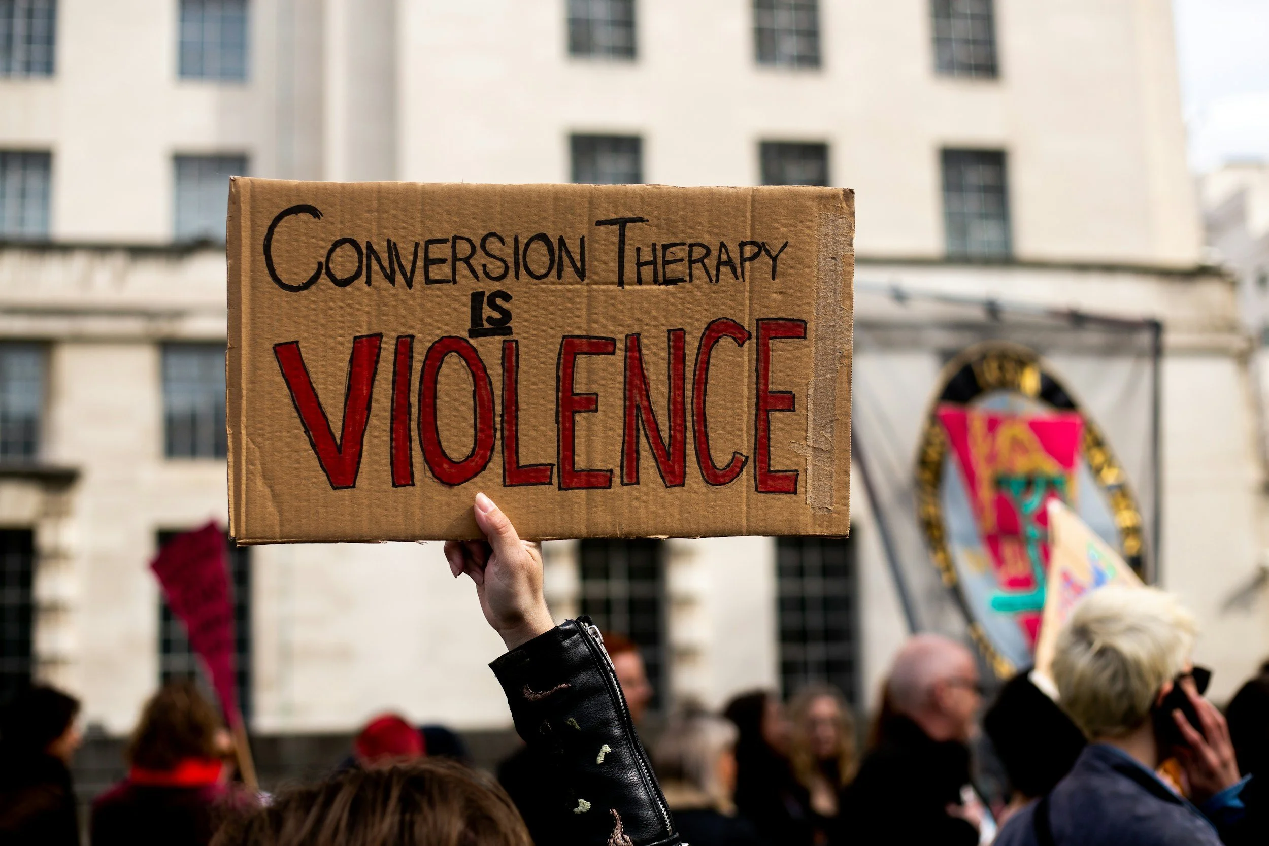 Person holding a cardboard protest sign that says "Conversion Therapy is Violence" during a demonstration with a crowd and buildings in the background.
