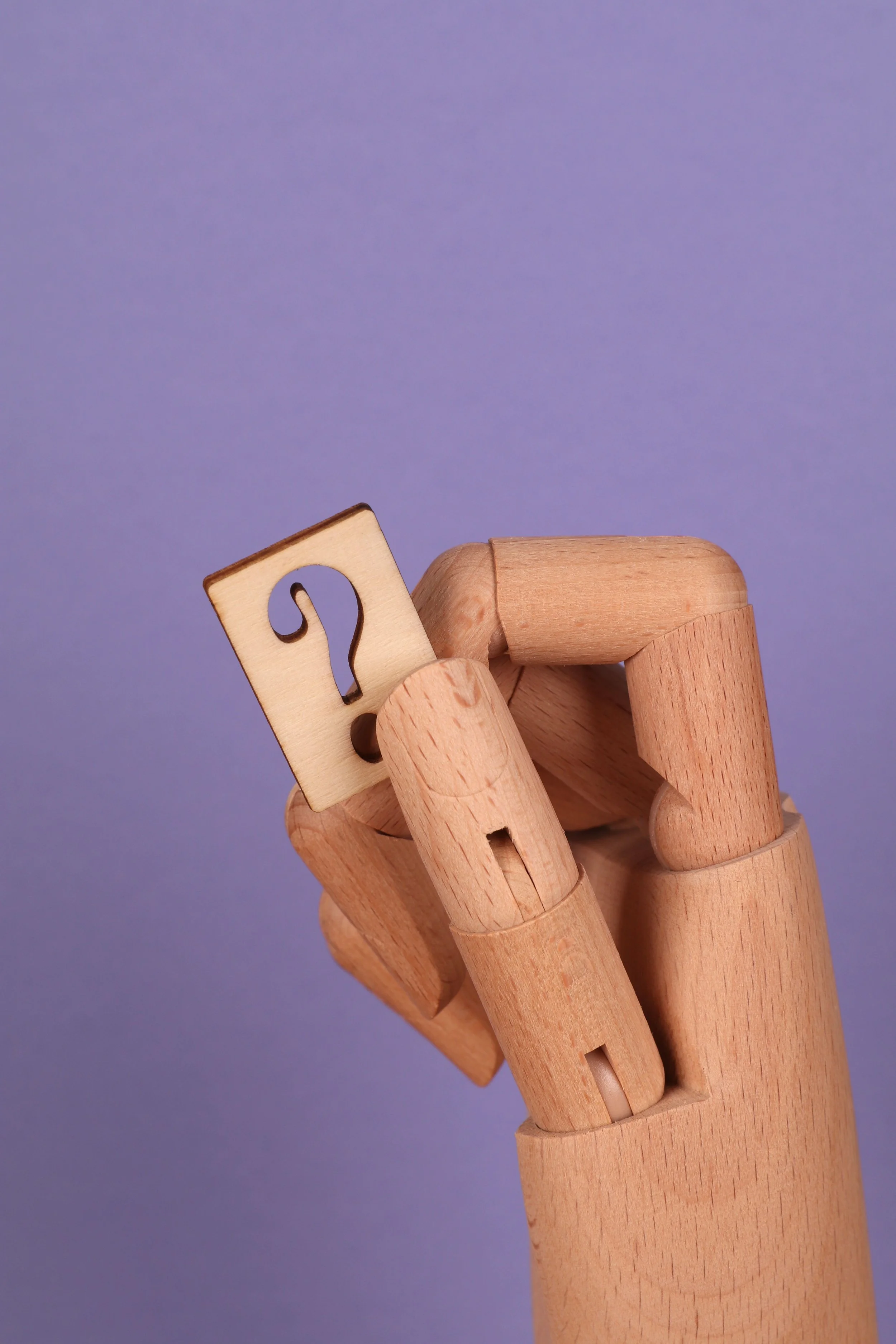 A wooden mannequin hand holding a block with a question mark on it, against a purple background.