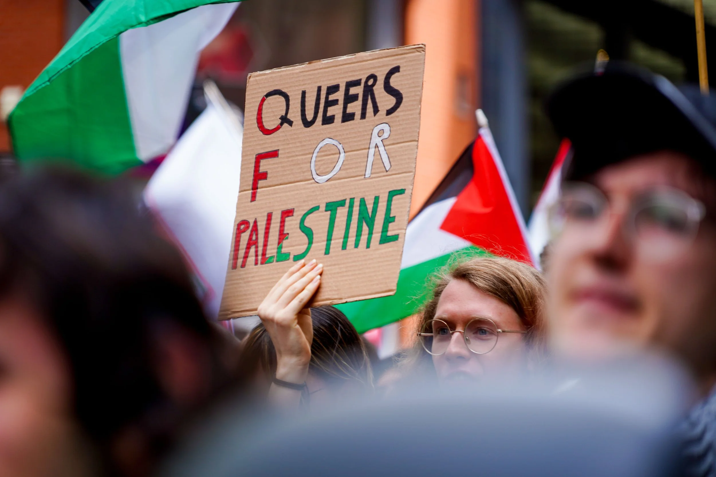 A crowd at a rally holding a cardboard sign that reads 'Queens for Palestine,' with numerous flags and umbrellas visible in the background.