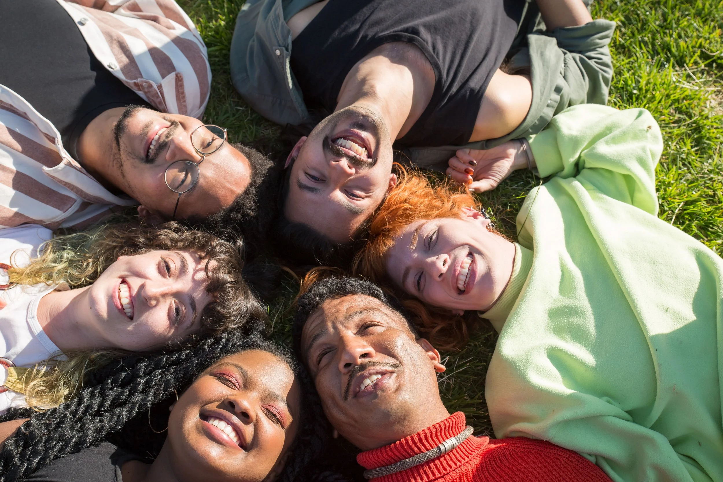 A group of diverse young adults is lying on the grass in a circle, smiling and enjoying each other's company on a sunny day.
