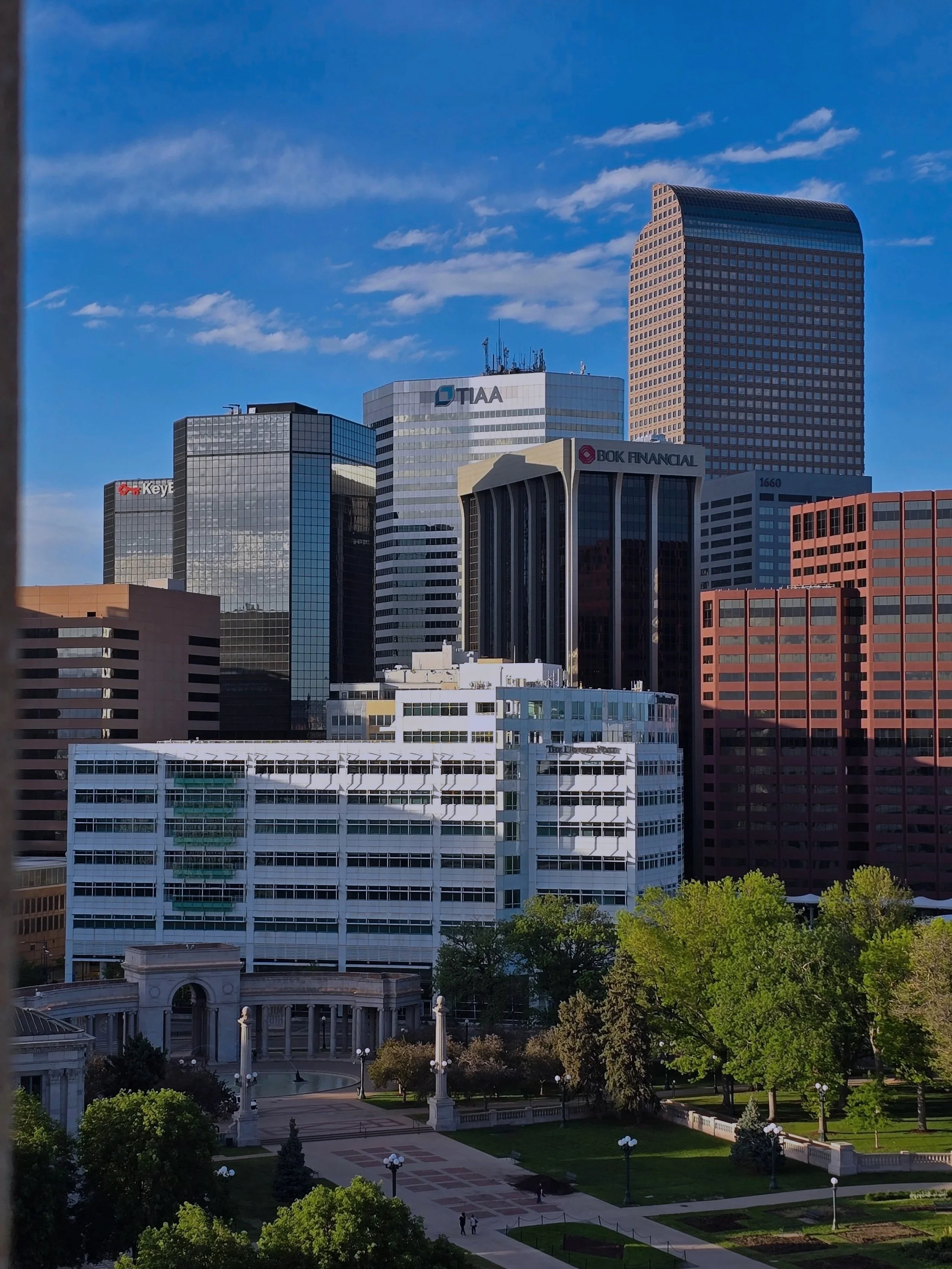 The Denver cityscape with tall modern skyscrapers against a blue sky with scattered clouds. The foreground features a plaza with trees, lampposts, and a fountain, surrounded by classical architecture.
