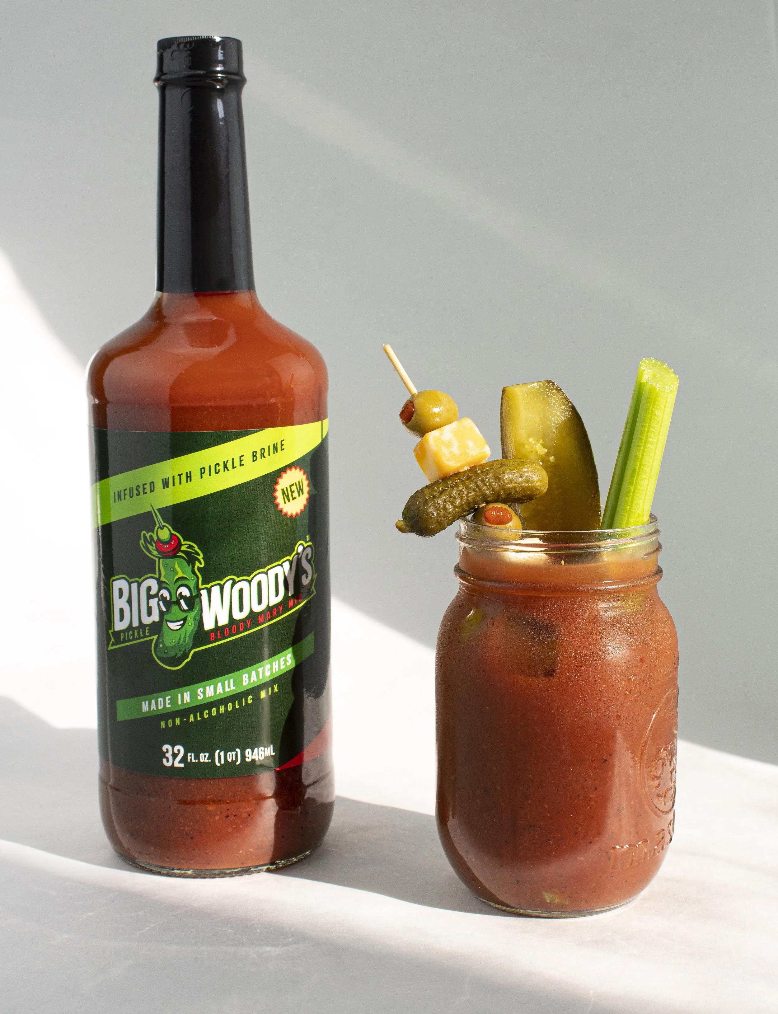 A bottle of Big Woody's spicy pickle brine, and a glass jar filled with a bloody mary garnished with a pickle spear, celery stalk, and skewered olives and cheese.