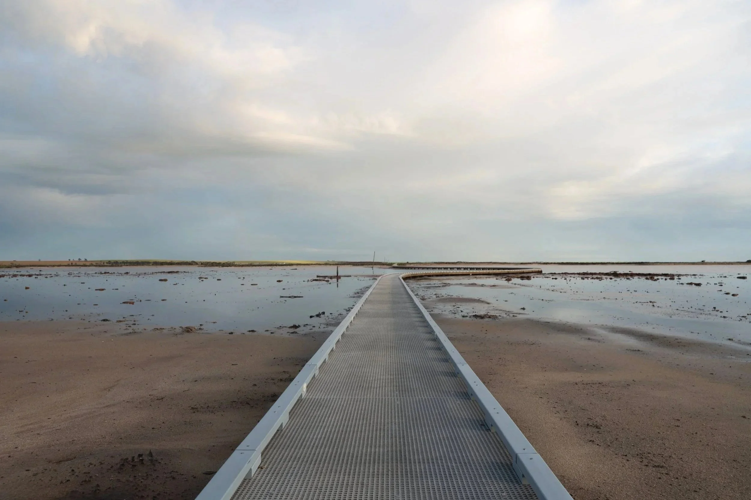 A metal walkway extends into a shallow body of water on a cloudy day with a cloudy sky.