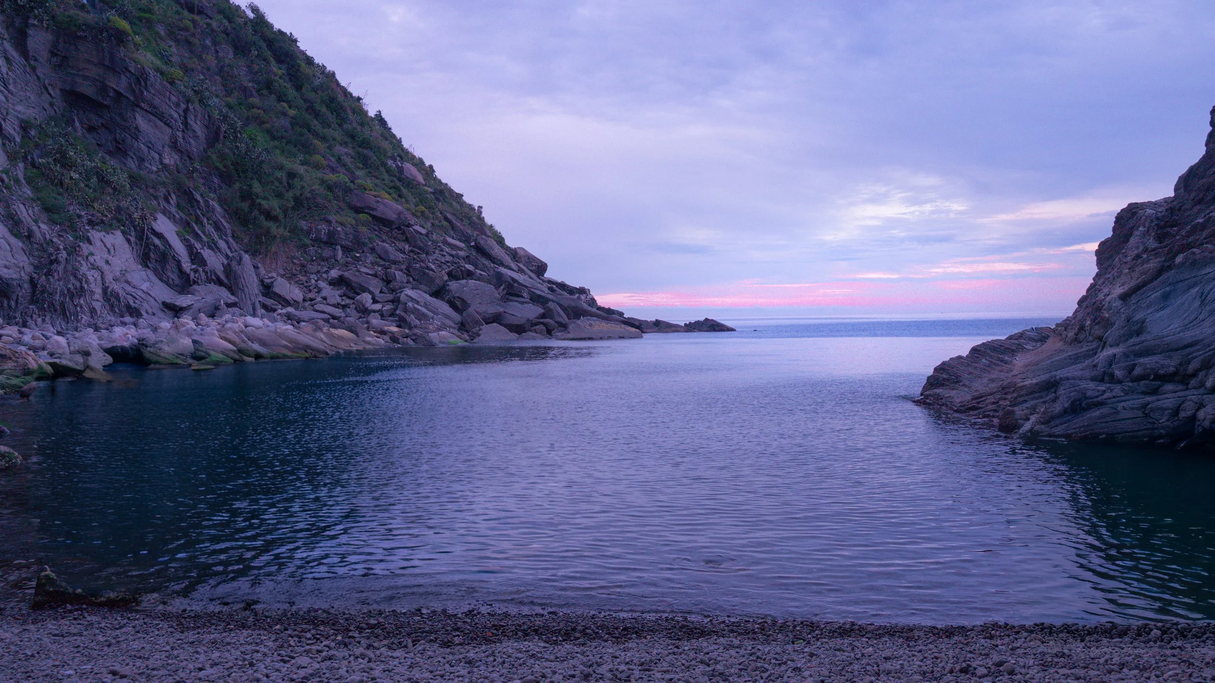 Sunrise over a tranquil cove with calm water surrounded by rocky hills and a pebble beach.