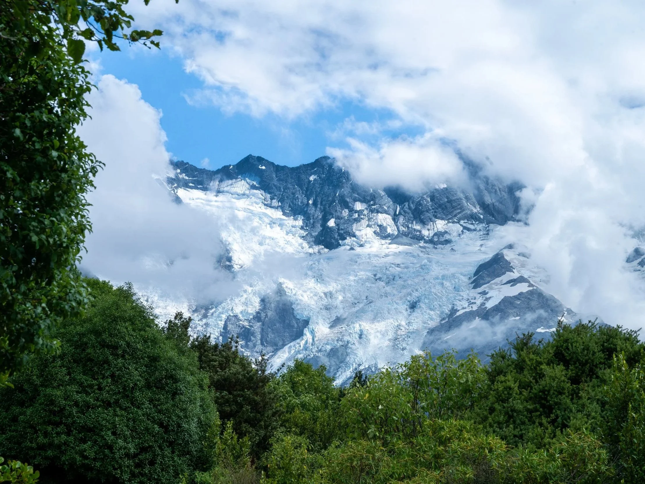 Snow-covered mountain with rocky peaks partly shrouded in clouds, surrounded by green trees and foliage in the foreground, under a blue sky with scattered clouds.
