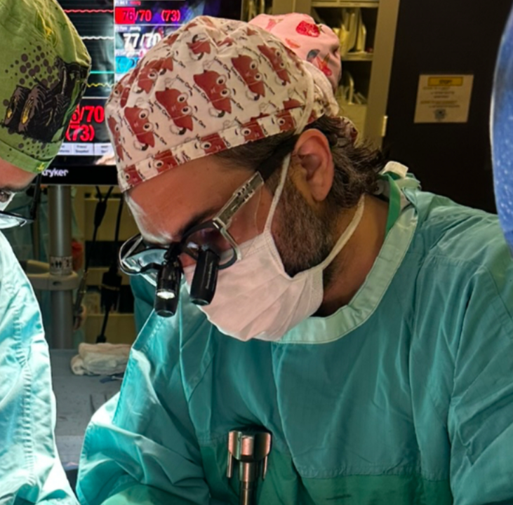 Surgeon in operating room wearing surgical mask, glasses with magnifying lenses, and a scrub cap with bear pattern, focused on a surgical procedure.