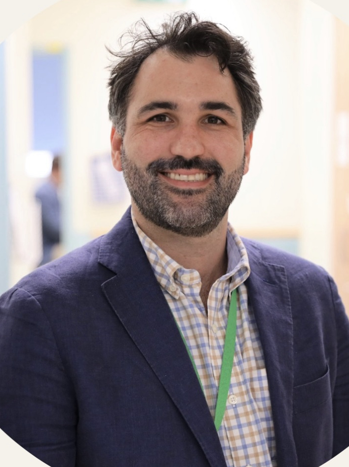 A smiling man with dark hair and a beard, dressed in a blue blazer and checked shirt, standing in a well-lit indoor setting.