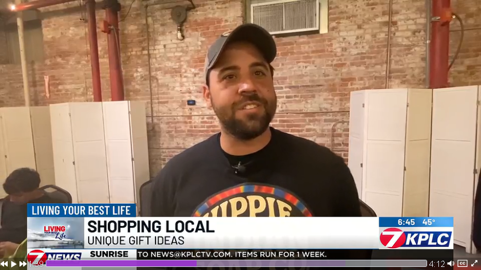 A man wearing a baseball cap and black T-shirt with colorful text stands in front of a brick wall with lockers behind him; news banner at the bottom promotes local shopping ideas.