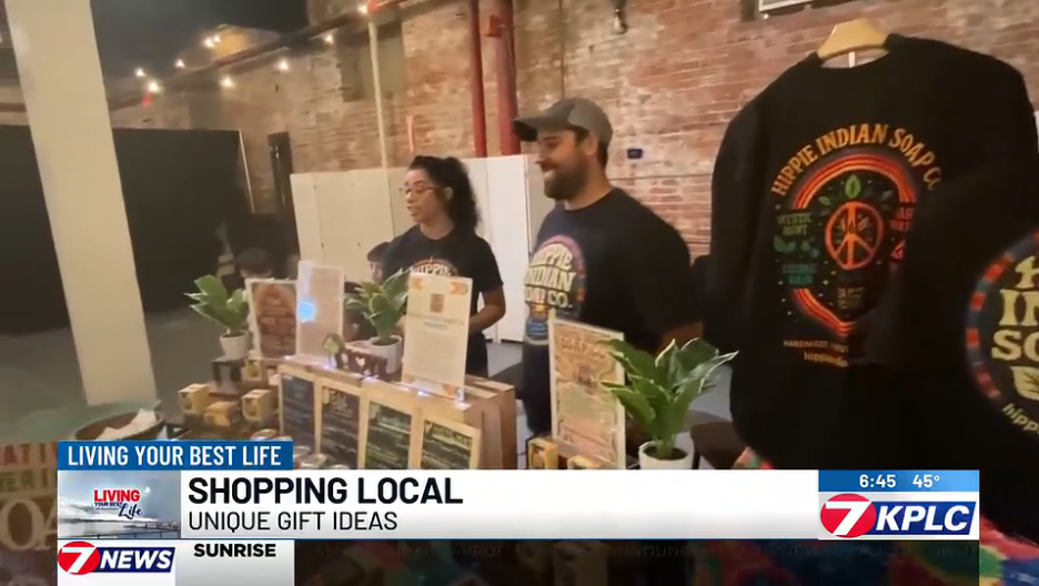 Two people standing behind a table selling soap and other products at a local market, with a brick wall in the background and a hoodie on display.