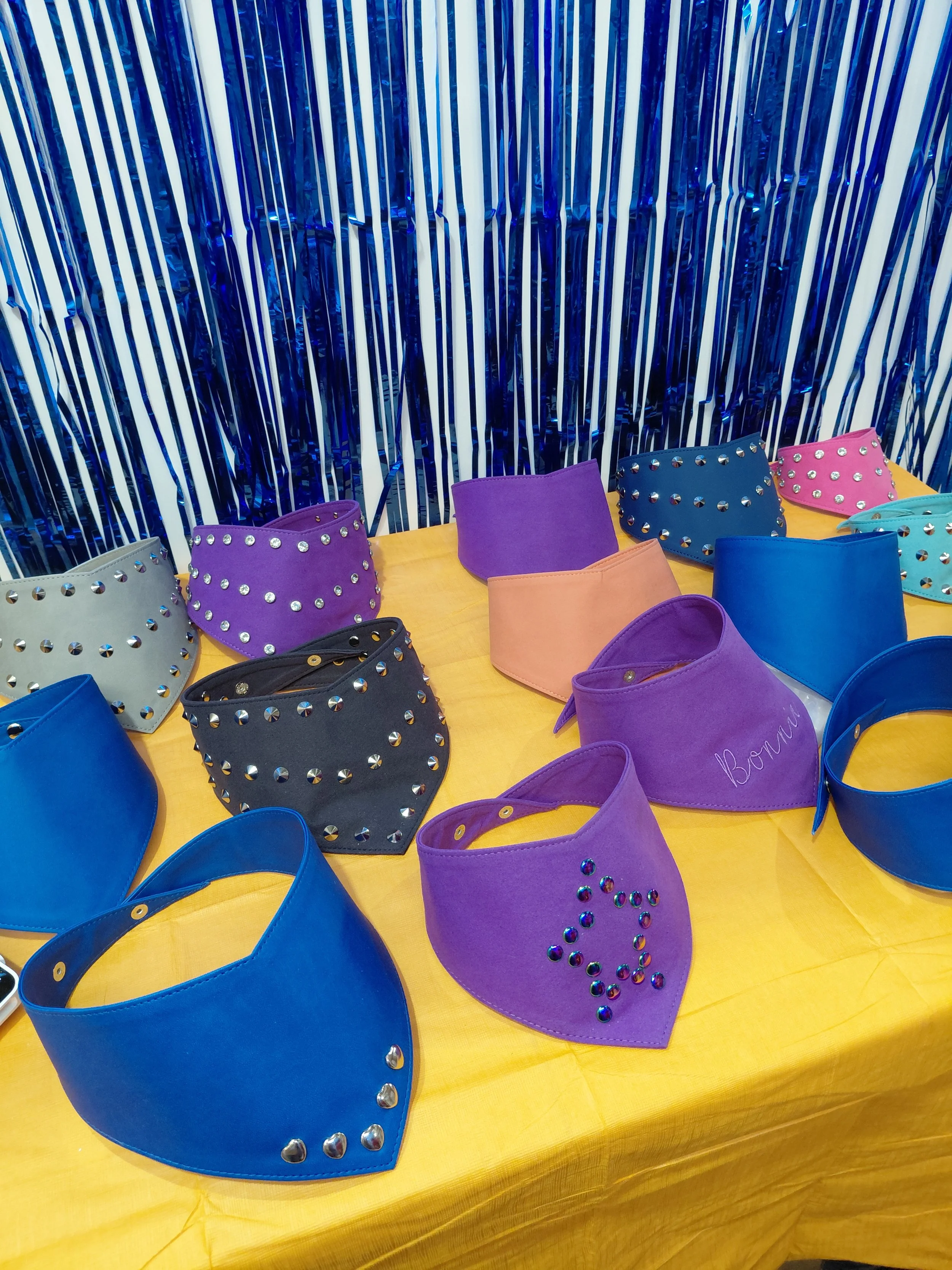 Colorful dog bandanas decorated with rhinestones displayed on a yellow table against a shiny blue and white striped backdrop.