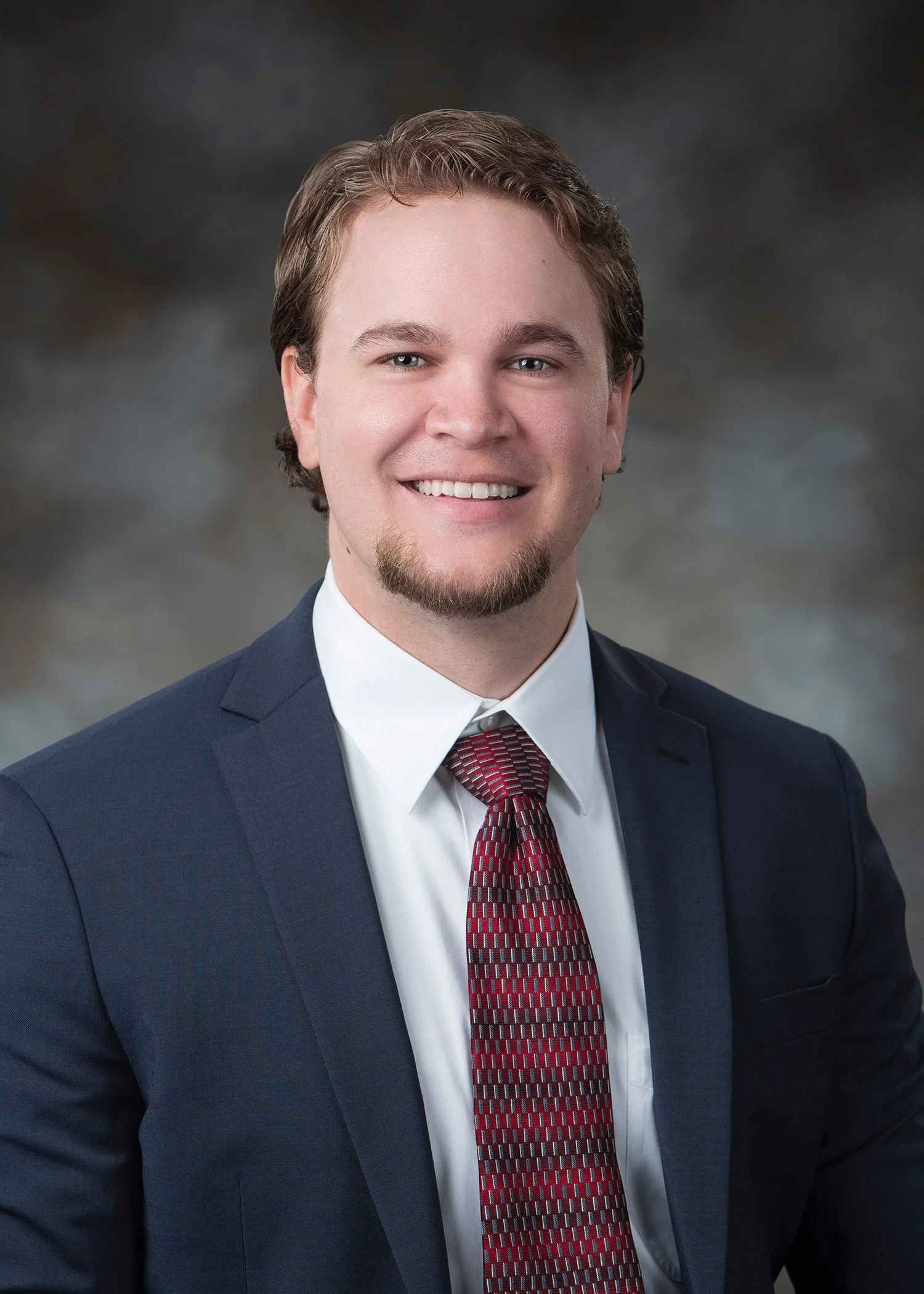 A man with light brown hair, blue eyes, and a goatee is smiling in a professional headshot. He's wearing a navy blue suit, a white shirt, and a red patterned tie, with a dark, blurred background.