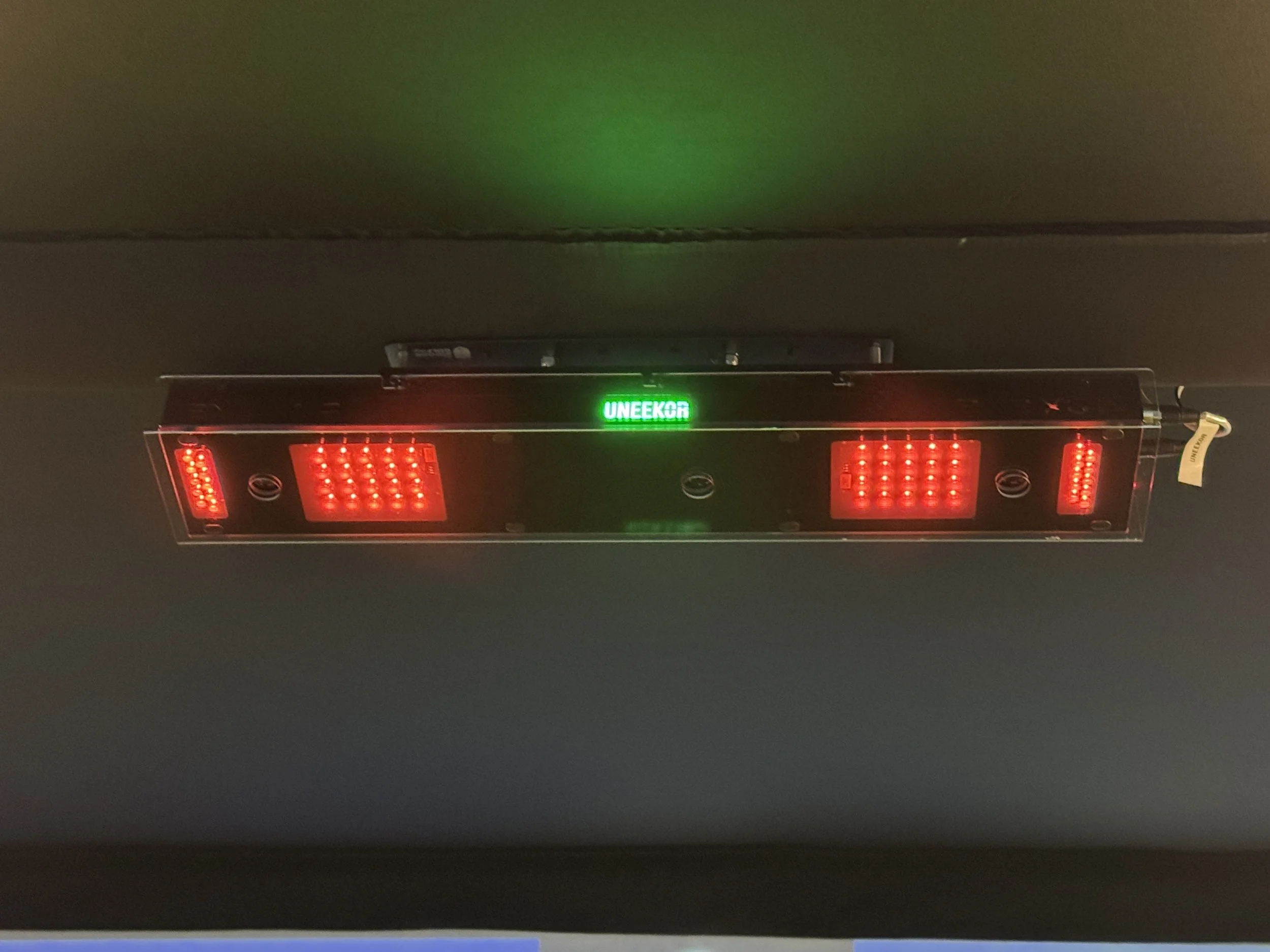A red LED traffic light system with multiple lights on a black panel and a green illuminated sign in the center that reads 'UNEKO'. The system is mounted on a ceiling.