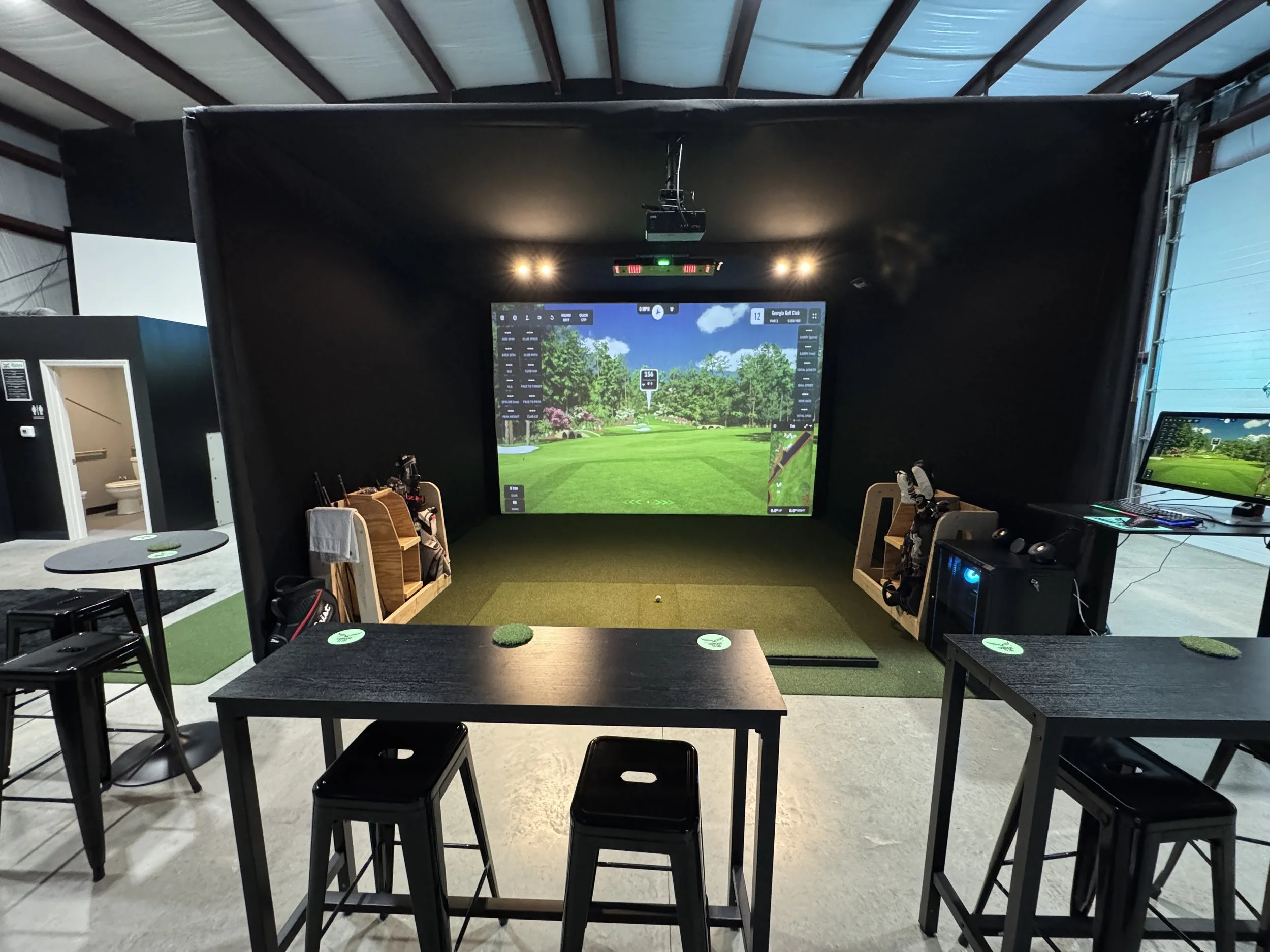 Indoor golf simulation room with a large screen displaying a virtual golf course, golf clubs on racks, and seating area with tables and stools.