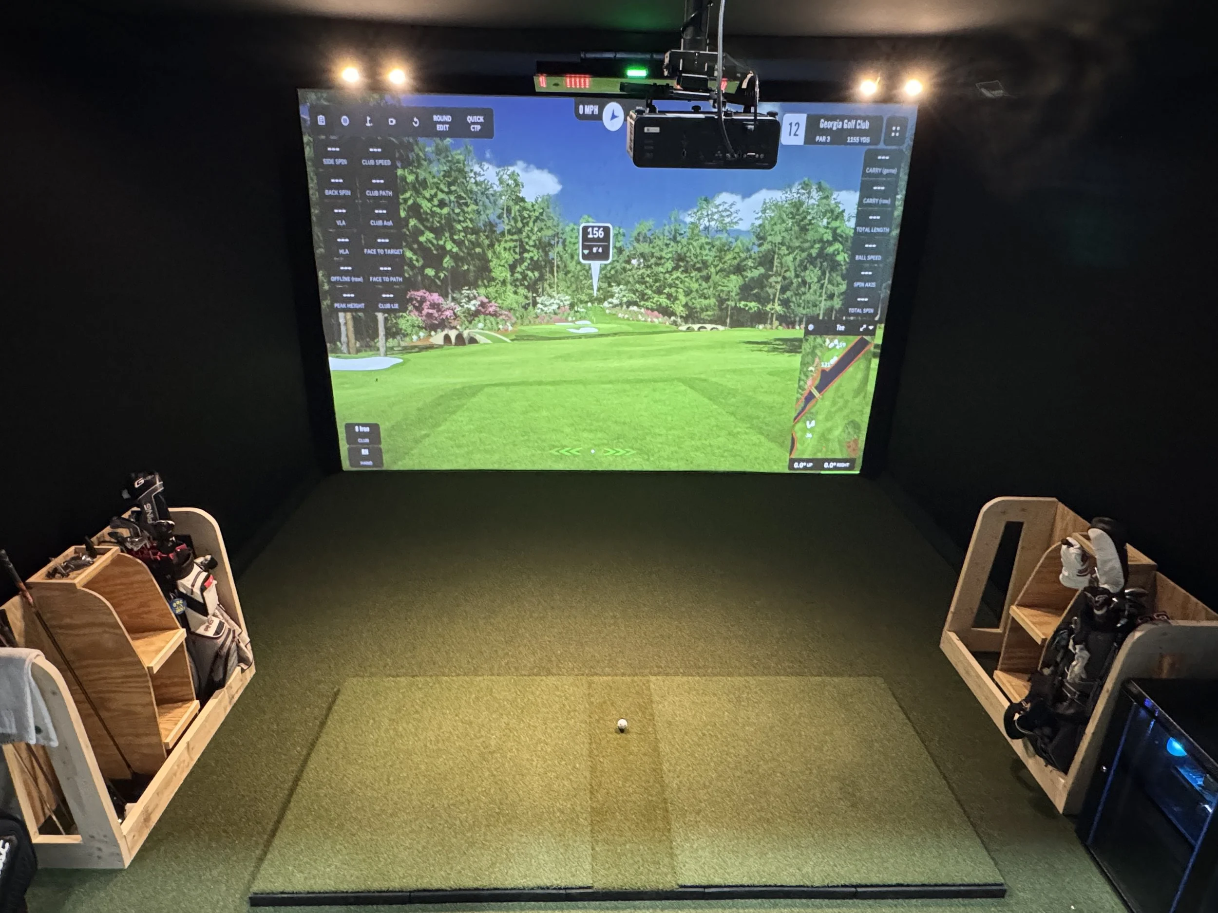 Indoor golf simulation room with a large screen displaying a golf course, golf clubs and bags on wooden racks, and a golf ball on the mat.