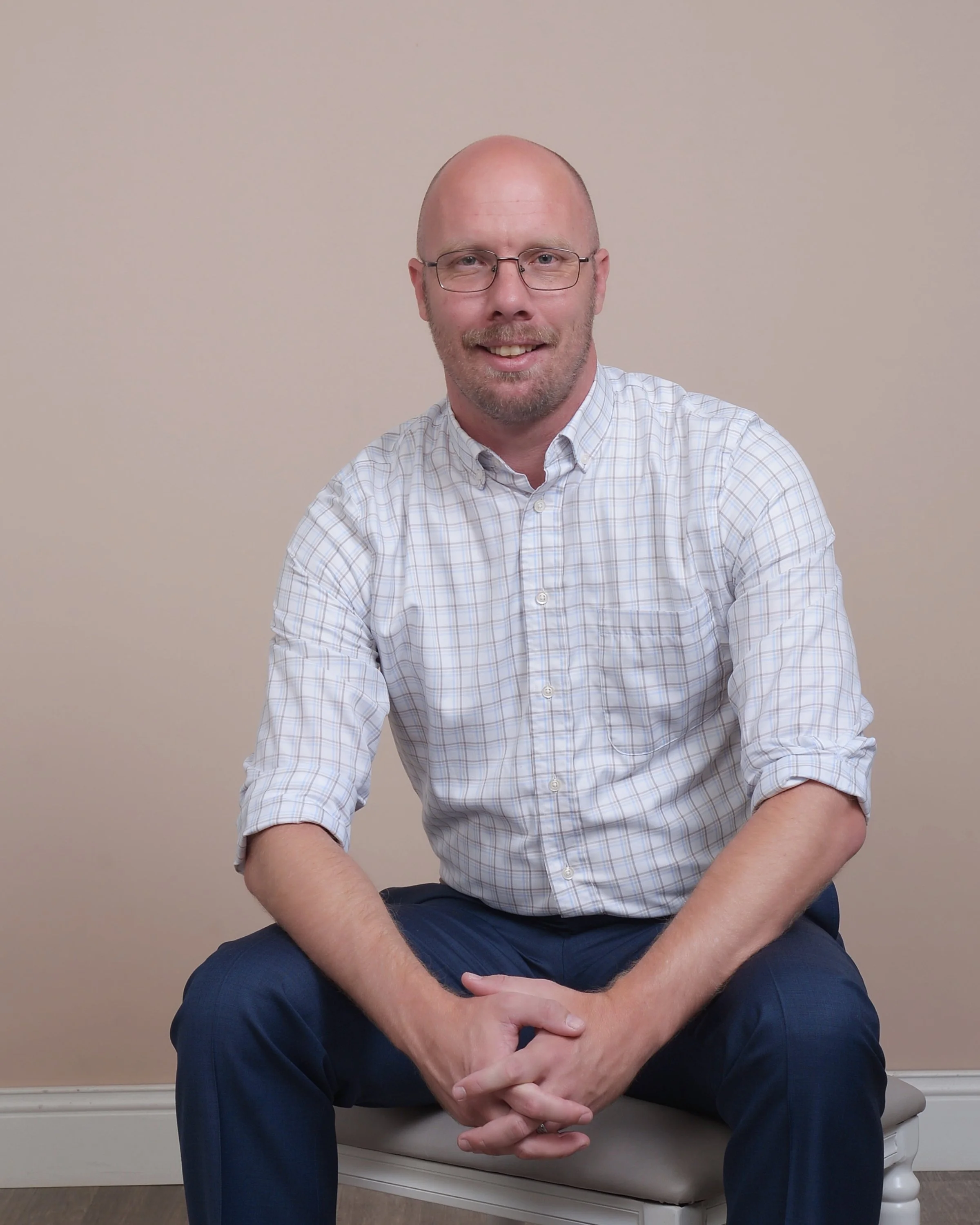 A man with glasses, a bald head, and a beard sitting on a beige bench, smiling, wearing a checked shirt and dark pants, against a plain beige background.