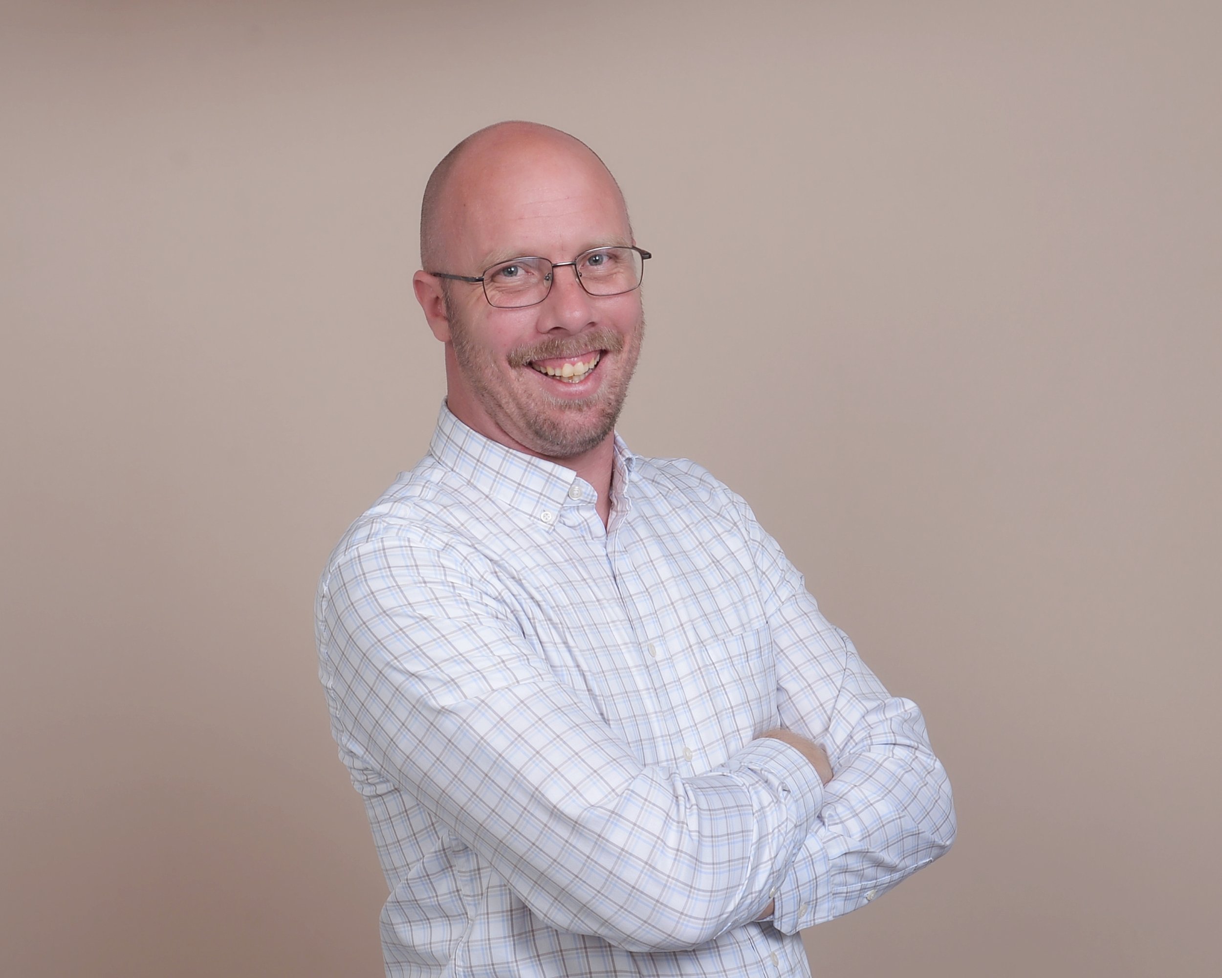 A man with glasses and a beard, smiling with arms crossed, wearing a light checkered shirt, standing against a plain beige background.