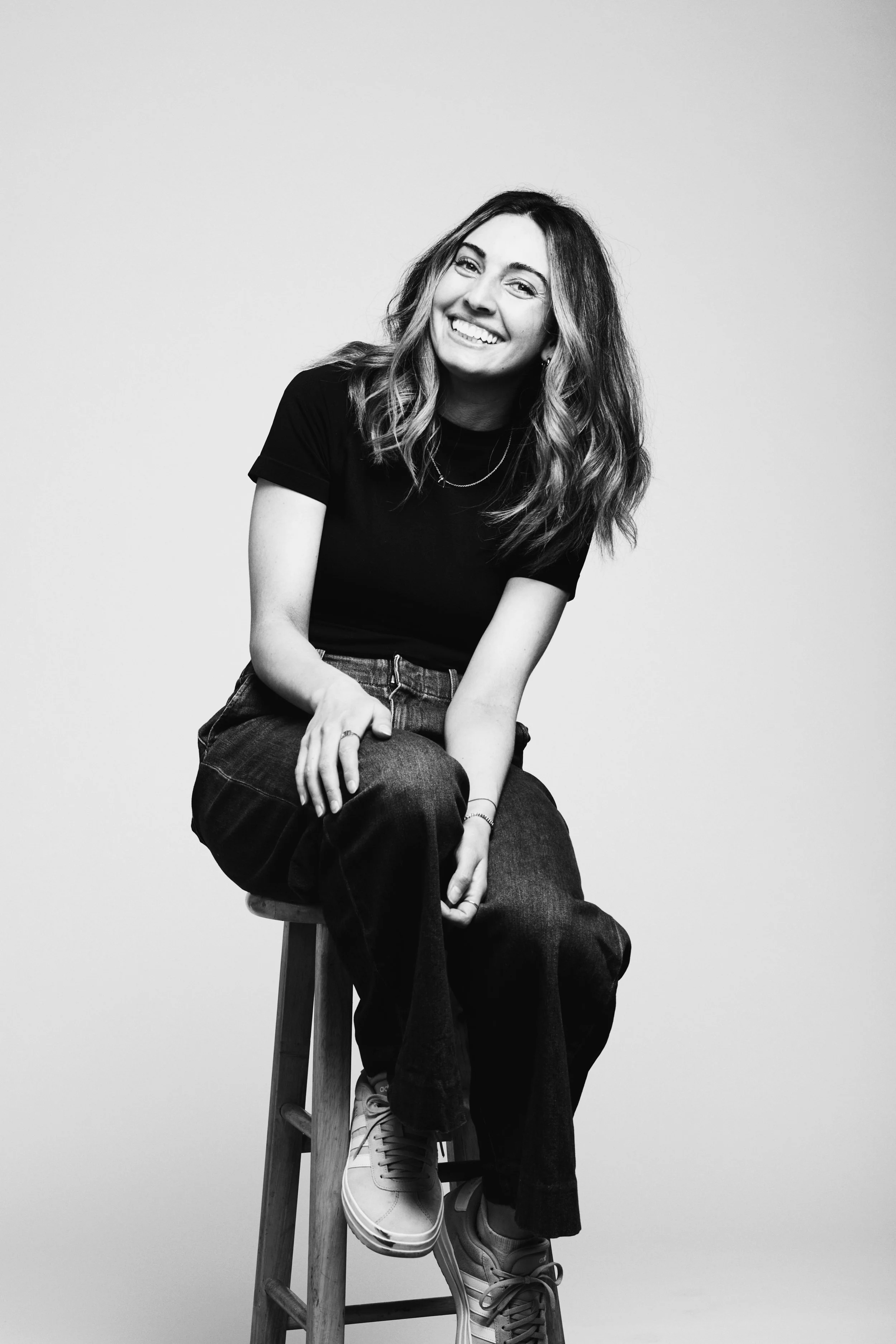 Black and white portrait of a woman sitting on a stool, smiling, with wavy hair, wearing a black t-shirt, jeans, and sneakers, against a plain background.