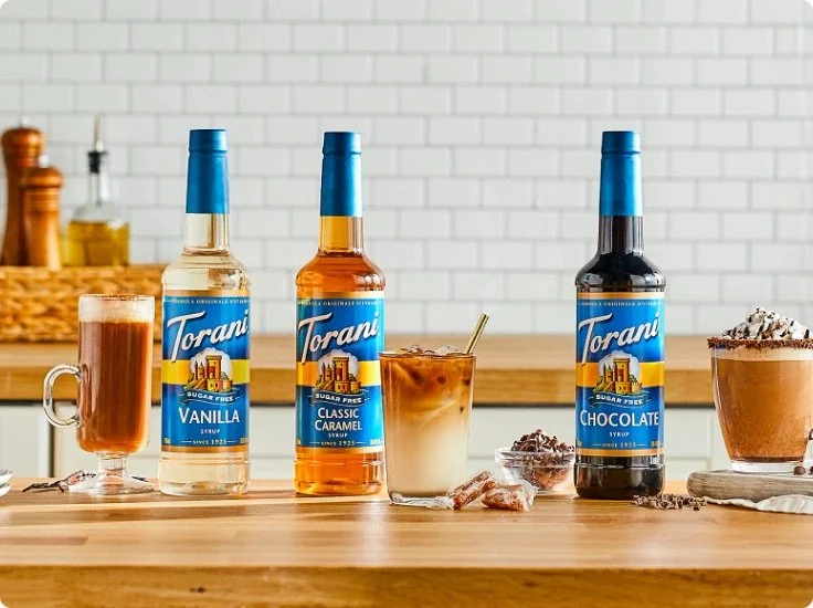 Three bottles of Torani syrup labeled vanilla, classic caramel, and chocolate are on a kitchen counter. There are drinks and dessert toppings nearby, including a chocolate drink in a mug, a layered coffee beverage, and a glass of hot chocolate. A bowl of whipped cream topped with chocolate shavings is on the right.