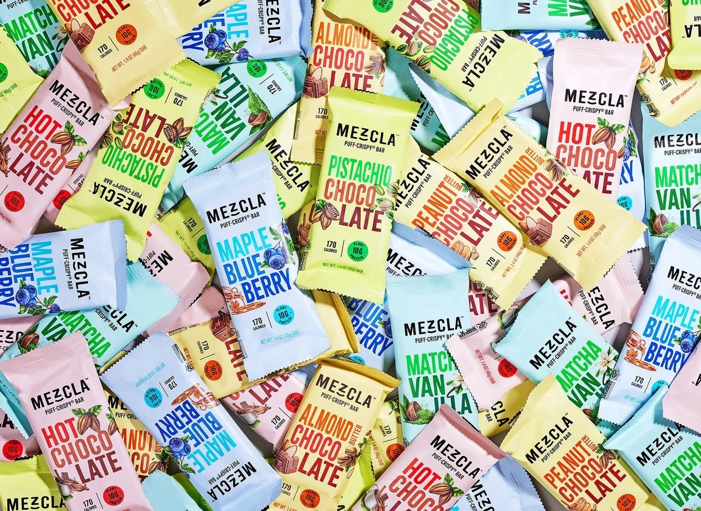 Assorted MEZCLA puff-crispy snack bars in colorful packaging including flavors such as Hot Chocolate, Almond Choco Latte, Pistachio Choco Latte, Maple Blueberry, and Matcha Vanila, scattered in a pile.