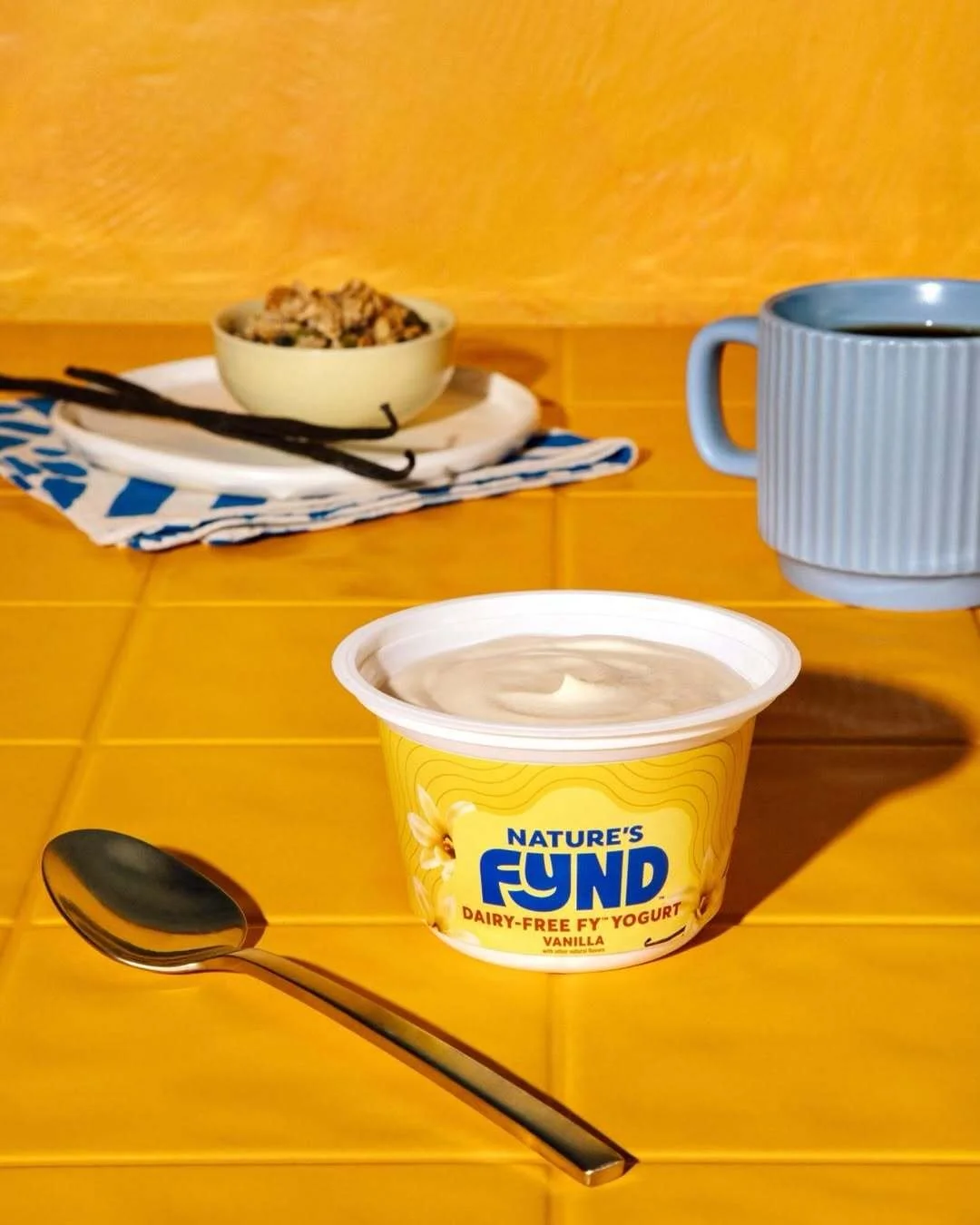 A container of Nature's Farm dairy-free vanilla yogurt, a metal spoon, a cup of black coffee in a blue striped mug, a bowl of granola with chopsticks on a white plate, all on a yellow tiled surface.