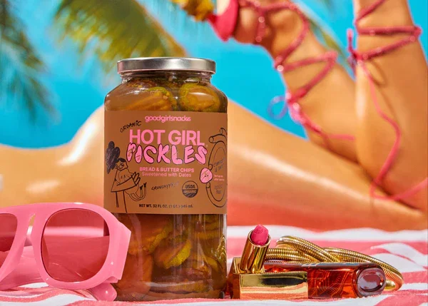 Two jars of hot girl pickles with colorful labels, garnished with herbs and peppers, are placed on a red background with ginger root, turmeric powder, and a honeycomb block on some miniature blue steps beside them.