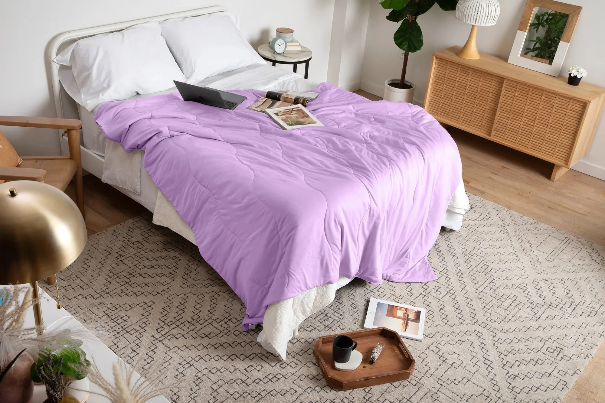 A bedroom with a bed covered in a purple comforter, a laptop, magazines, and a tablet on the bed, a side table with a clock, a large plant, a wooden cabinet with a mirror and a white table lamp, a beige rug, and a wooden tray with a black mug and a pen on the floor.