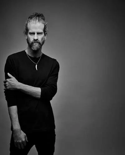 A man with long, wavy hair and a beard looking down, wearing a black t-shirt and a necklace with a pendant, against a plain white background.