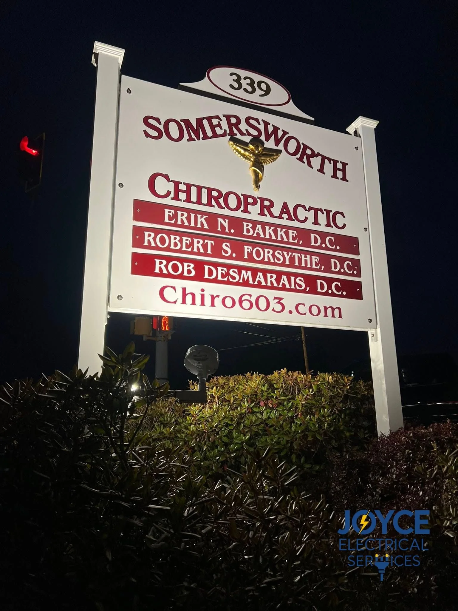 Sign for Somersworth Chiropractic featuring a gold caduceus symbol, list of chiropractors, and website, illuminated at night with surrounding foliage and streetlights.