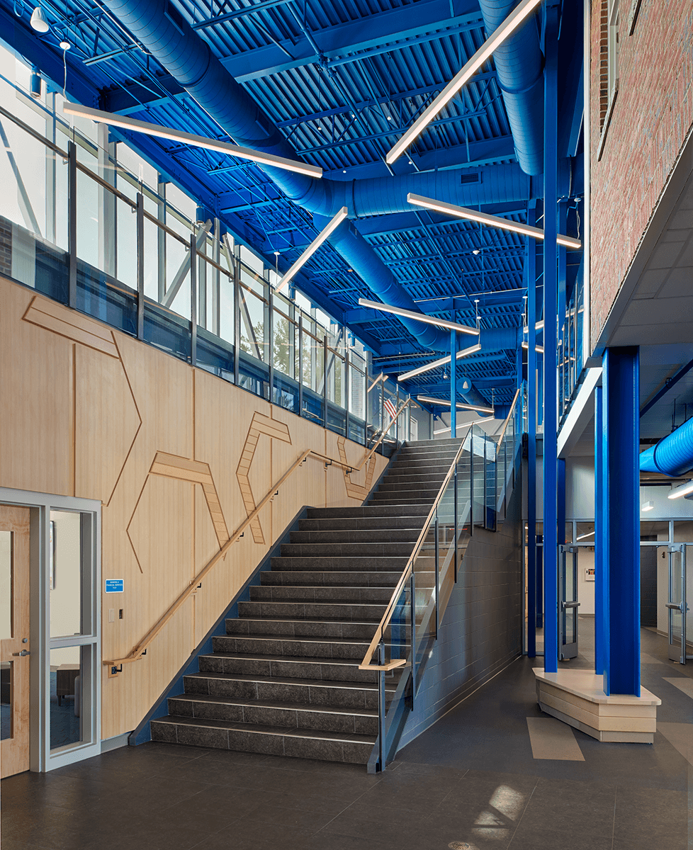 Modern indoor staircase with black steps, glass handrails, blue-painted structural elements, and a blue industrial ceiling with exposed ductwork and contemporary light fixtures.