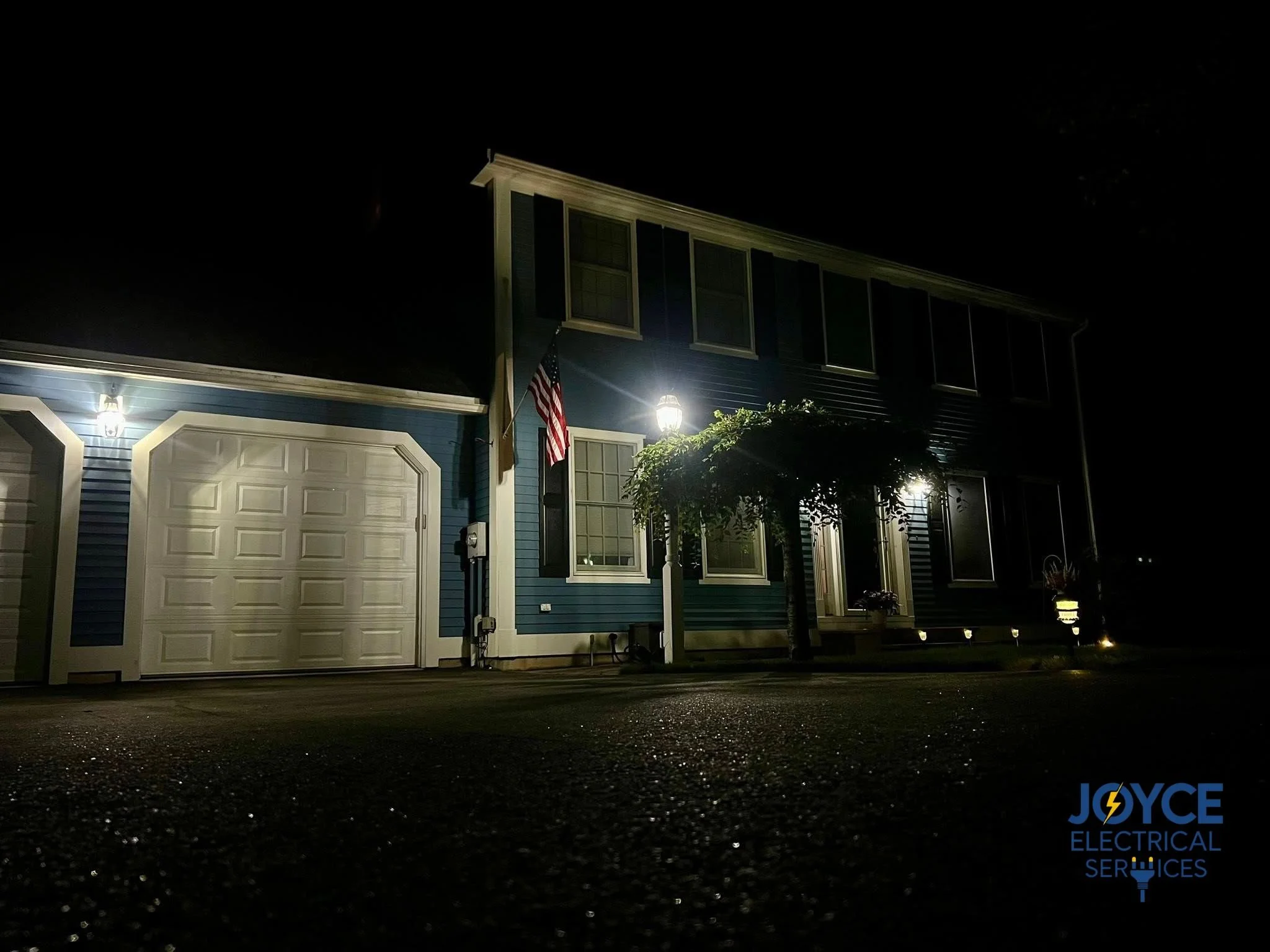 A house at night with exterior lighting, an American flag, and a sign for Joyce Electrical Services illuminated in front.