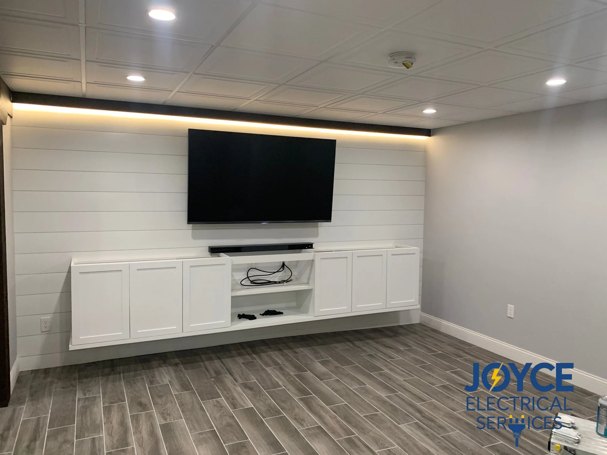 Empty room with a wall-mount television, white cabinet, and wood-look tile flooring, illuminated by ceiling and strip lighting.