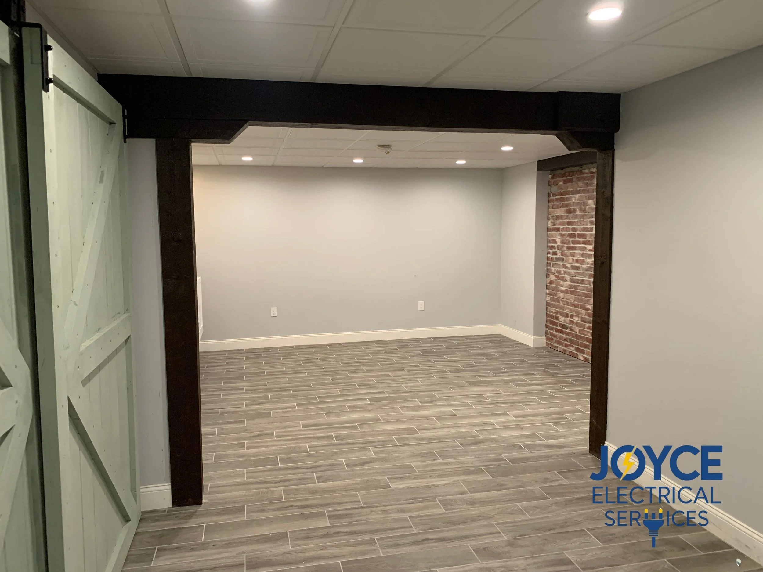 Empty room with wood-look flooring, grey walls, exposed brick wall section, ceiling with recessed lighting, and a decorative dark wood beam. A green sliding door is partially open on the left, and a logo for Joyce Electrical Services is in the bottom right corner.