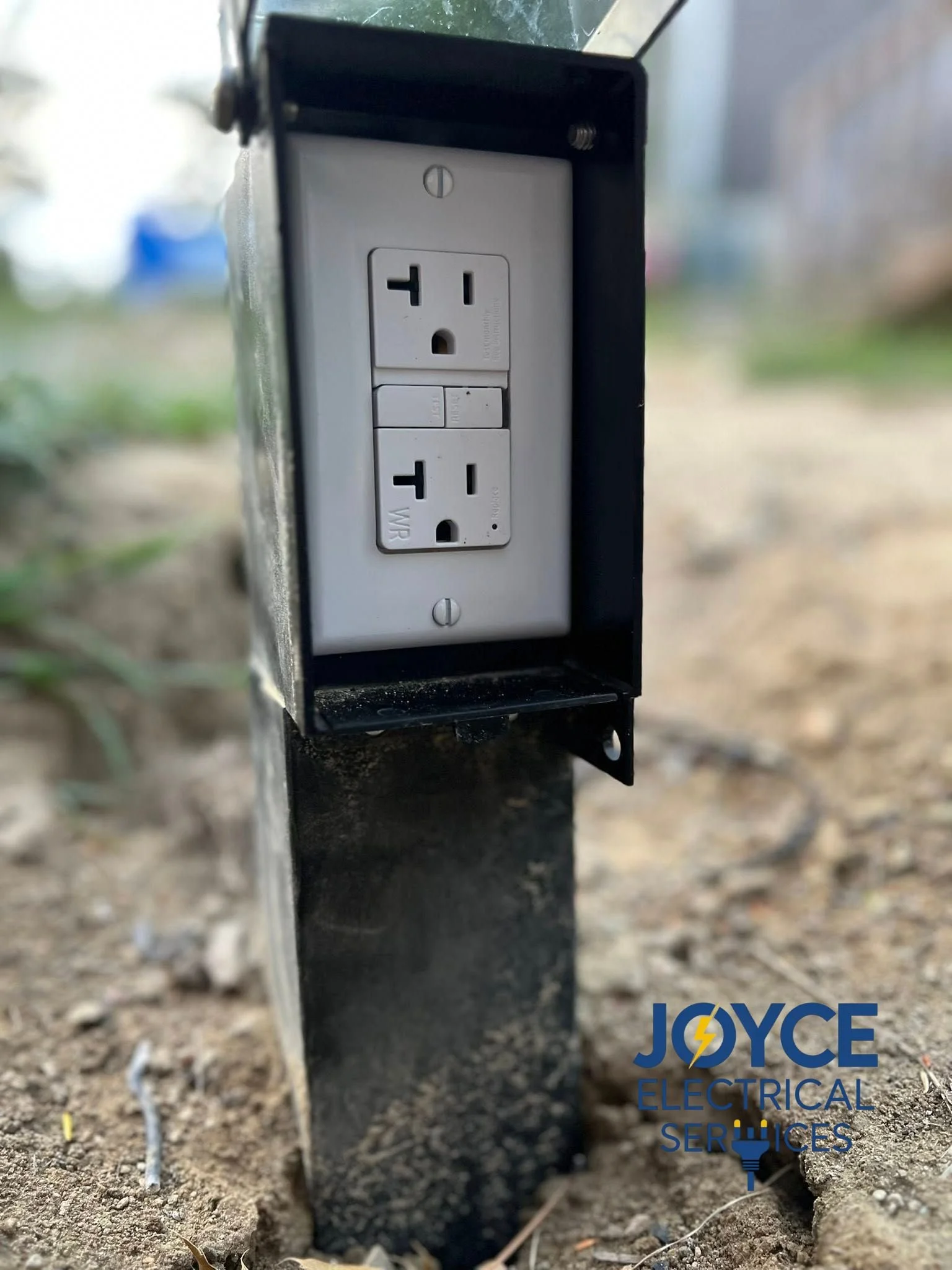 Close-up of an outdoor electrical outlet box with two power outlets, mounted on a black metal post, surrounded by dirt.
