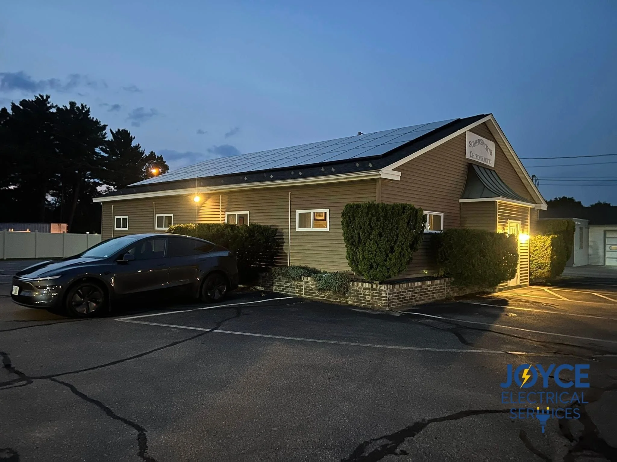 Parking lot with a black Tesla vehicle parked in front of a beige building with a sign that reads 'Somerworth Chiropractic.' The building has a solar panel roof and is illuminated by exterior lights; night scene with visible trees and power lines in the background.